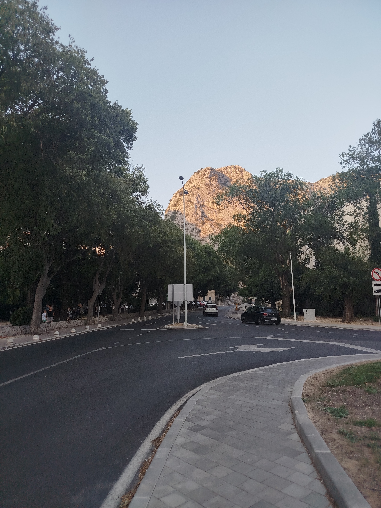 The image captures a street scene in Omiš, Croatia, with a prominent mountain range in the background. In the foreground, a paved sidewalk curves to the right, bordering a dark asphalt road. The road shows lane markings, including a directional arrow. Several cars are visible on the road, including a black hatchback and a white sedan.
A line of trees with dense green foliage flanks the left side of the road, with a few people walking along the sidewalk in the distance. White bollards line the edge of the sidewalk. A street lamp is positioned on the right side of the road.
The mountain range in the background is rugged and appears to be illuminated by sunlight, casting shadows. The sky is clear and pale blue. On the right side of the image, a traffic sign with a red circle and a diagonal bar is visible, indicating a prohibition, along with a symbol of a truck. Another signpost is also present. The overall scene suggests a moderately busy urban environment adjacent to natural scenery.