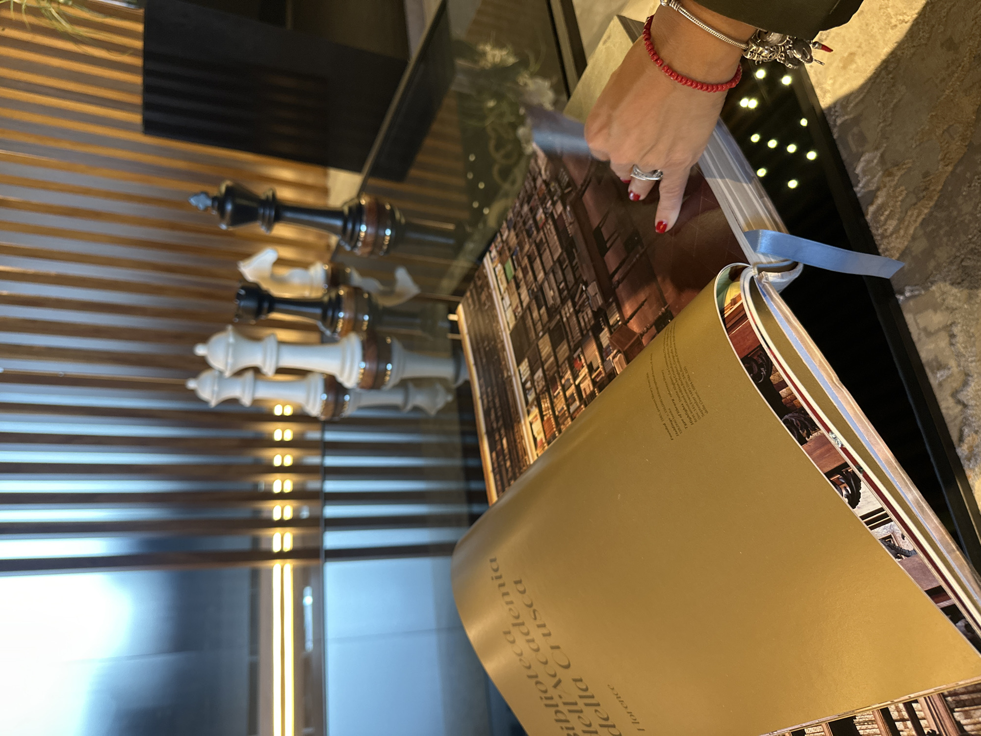 The image shows a partially opened book on a glossy surface, possibly a coffee table or counter, with a person's hand resting on the open page. The book has a gold-colored cover with the text "biblioteca academia della Crusca" visible. The pages within the book contain images of what appear to be library interiors. A blue ribbon bookmark is visible. Adjacent to the book, on the left side of the image, are several chess pieces. The surface the book and chess pieces rest on is reflective, mirroring the chess pieces and a backdrop of horizontal striped patterns that suggest a wall or a decorative panel. The environment seems to be an interior space, possibly a lounge or a study area.