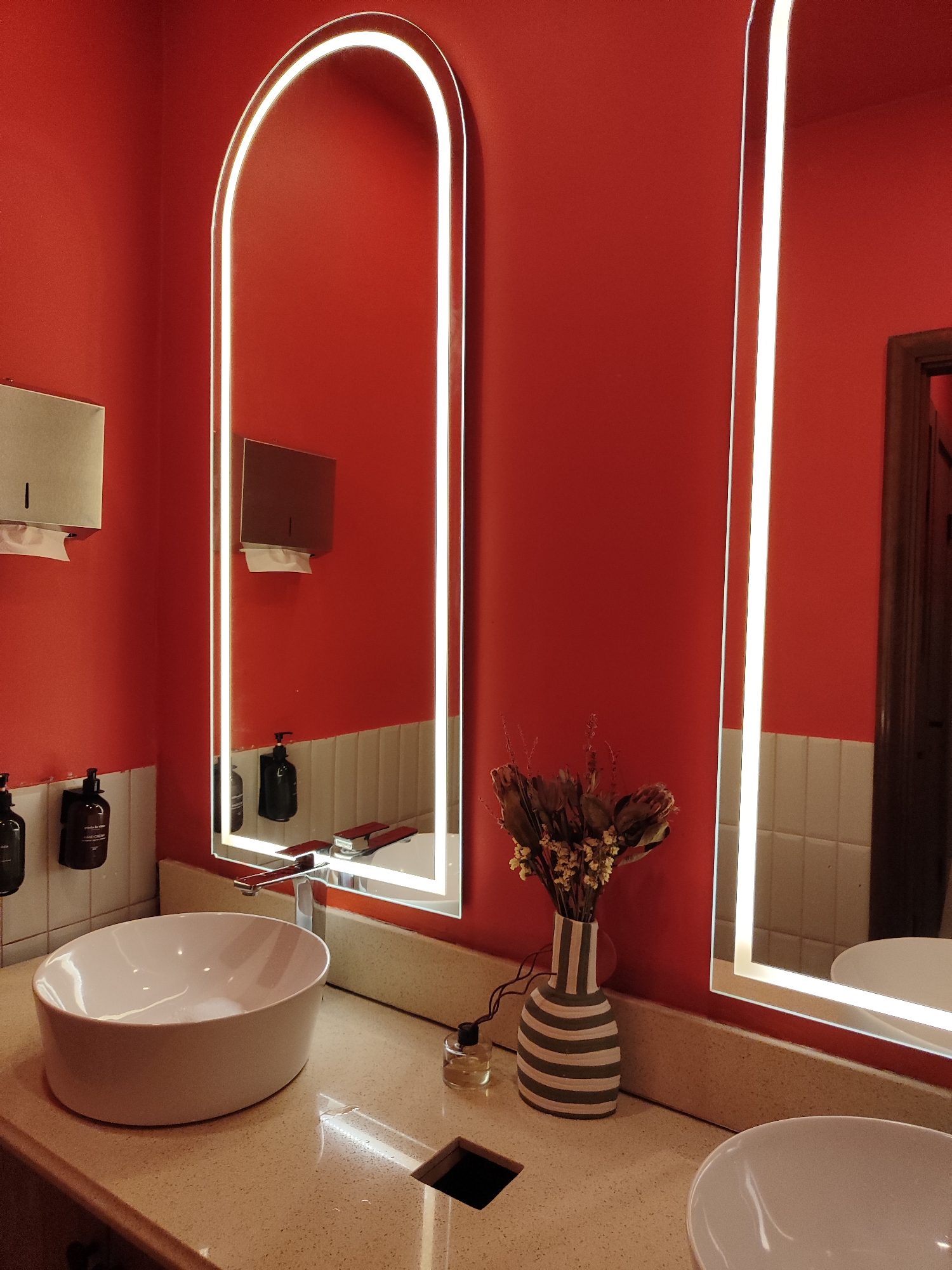 This is a photograph of a modern bathroom with red walls. On the left side of the image, a large arched mirror with integrated lighting is visible, reflecting a dispenser on the wall above a sink. Below the dispenser, two bottles of hand soap are mounted on the wall. To the right of this, a second, rectangular mirror with integrated lighting is present. This mirror reflects a sink, a faucet, and a vase of dried flowers on the countertop. The countertop is speckled and appears to be made of stone or quartz. In the foreground, a white ceramic sink is situated on the countertop. Another sink is partially visible in the far right of the image. The overall scene suggests a clean and stylish public restroom, potentially in a restaurant, hotel, or shopping center in Almaty, Kazakhstan.