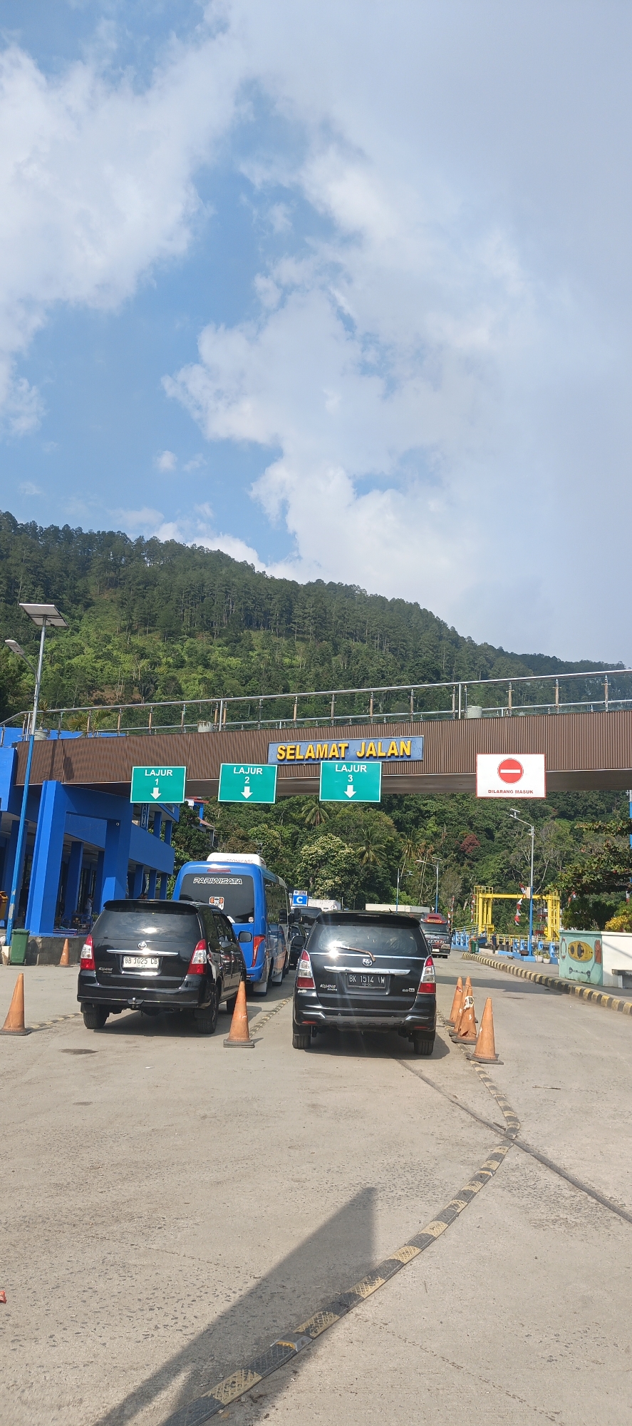 The image shows a view of a toll plaza on a sunny day with a mountainous, forested landscape in the background. The sky is blue with scattered white clouds. In the foreground, there are several cars queuing at the toll booths. The toll plaza itself is an elevated structure with blue and brown elements. Above the lanes, there are signs with the text "SELAMAT JALAN" (meaning "Have a nice trip" in Indonesian) and green signs indicating "LAJUR 1", "LAJUR 2", and "LAJUR 3" (meaning "Lane 1", "Lane 2", and "Lane 3"). There is also a red and white prohibition sign with a red circle and a horizontal bar, indicating "DILARANG MASUK" (meaning "No Entry"). Orange traffic cones are placed along the lanes. A solar panel is visible on a pole to the left of the toll plaza. The road surface is asphalt and has speed bumps. The cars appear to be modern passenger vehicles. The overall scene suggests a typical traffic situation at a toll collection point in Indonesia. The location is specified as Nagaritongah, Indonesia.
