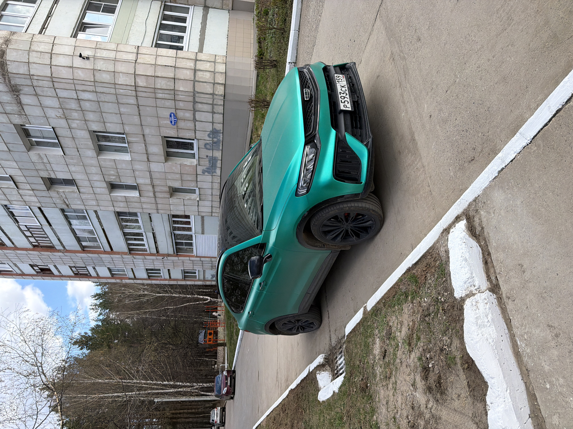 This image captures a vibrant teal/mint green SUV, likely a Geely Coolray/Binyue, parked on a paved surface in a residential area of Chaikovsky, Russia, during daytime. The car's front faces left, prominently displaying its grille with the Geely logo and a Russian license plate "P593CK 159 RUS." The "159" regional code corresponds to Perm Krai, where Chaikovsky is located.
In the background, a multi-story concrete apartment building dominates the left side of the frame, featuring numerous windows. A blue oval street sign is affixed to the building's facade, clearly showing the Russian word "Сосновая" (Sosnovaya), which translates to "Pine Street" or "Forest Street."
Beyond the parked car and the building, a natural landscape of mostly bare deciduous trees, possibly birches, and some evergreen trees stretches towards the horizon, suggesting an early spring setting. The sky above is partly cloudy. The foreground features a strip of sparse grass and dirt separated by a white-painted concrete curb. Other parked cars are visible further down the street in the distance. No people or specific activities are depicted, portraying a static scene of an urban environment.