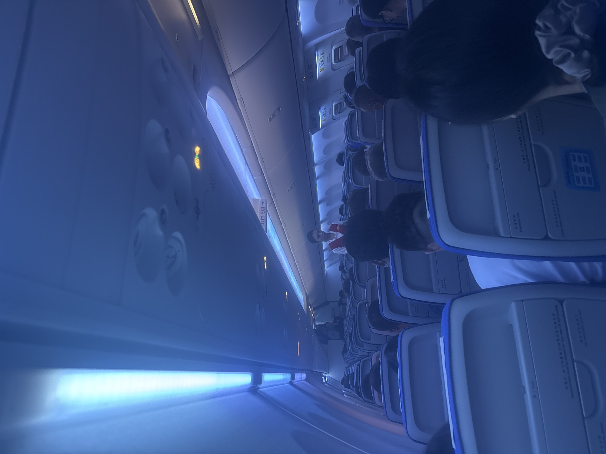 The image depicts the interior of an airplane cabin, likely during a flight, as indicated by the presence of rows of seats, overhead compartments, and cabin crew members. The lighting is predominantly blue, possibly due to ambient lighting or filters. Several passengers are seated, facing forward, with some partially visible. Two cabin crew members are visible; one standing near the front of the cabin, and another walking down the aisle. Signs, including "EXIT" signs in Chinese and English, are visible above the seats, suggesting this is an international flight or a flight with international signage standards. The overall scene is consistent with a typical airplane cabin environment, with no specific identifiable landmarks that would pinpoint its location within Xinwu District, China, or any other city. The image is taken from a seated passenger's perspective, looking towards the front of the cabin.