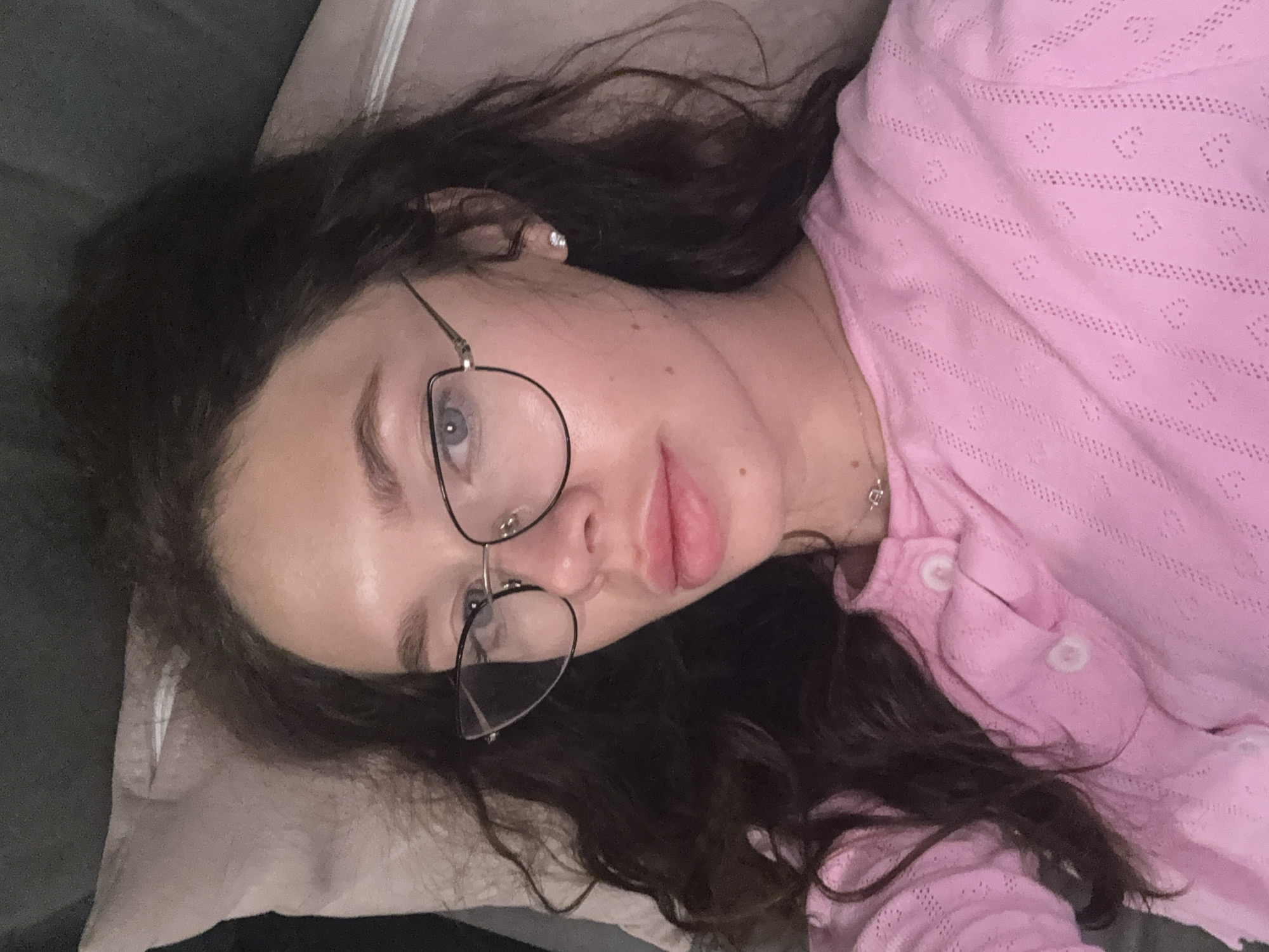 A woman is lying down, likely on a pillow or cushion. She is wearing eyeglasses with thin, black frames and a pink, buttoned shirt with a textured pattern. Her dark hair is loose and spread around her head. She appears to be indoors. No specific indication of Tel-Aviv, Israel.
