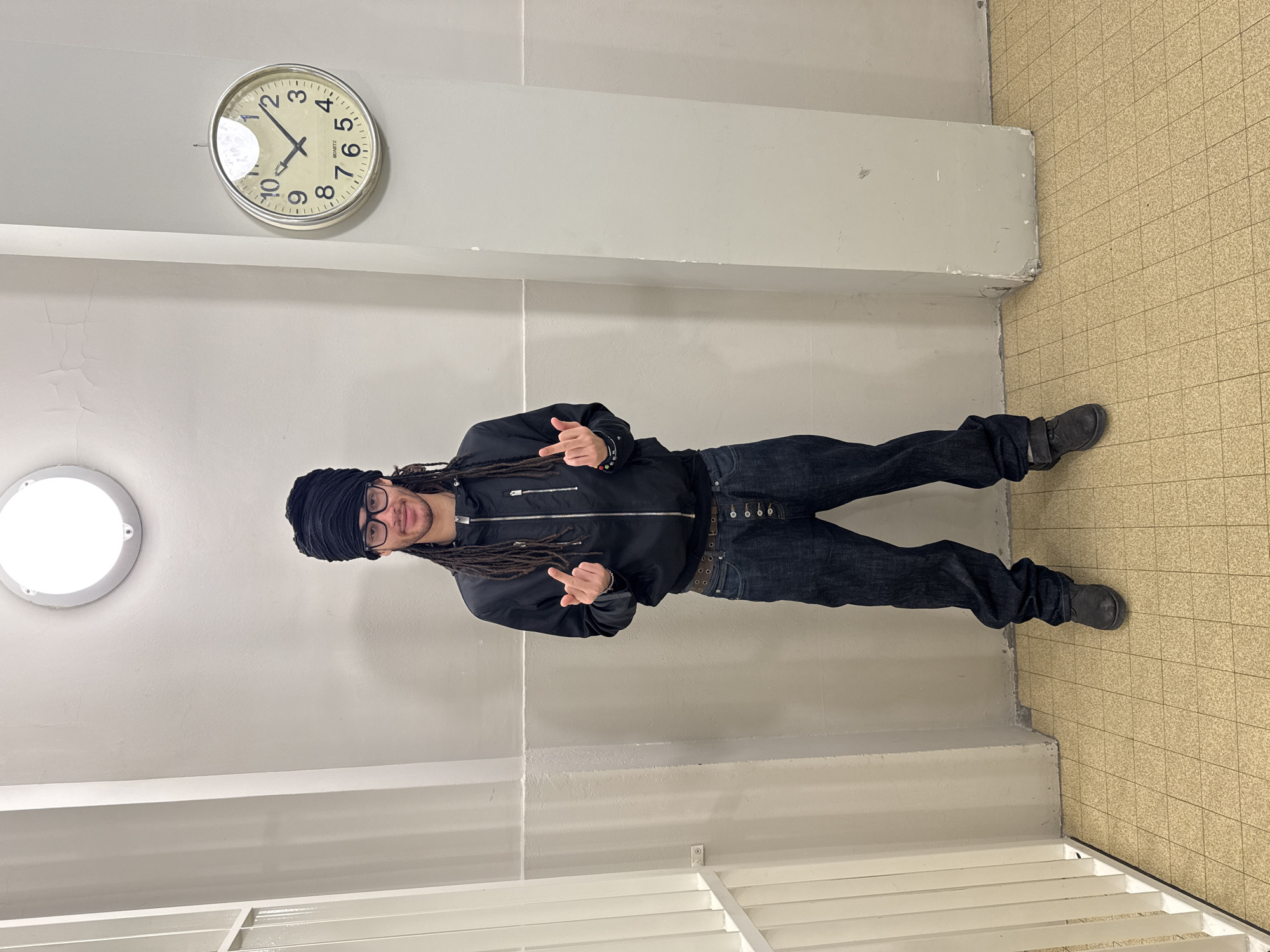 A person with dreadlocks, wearing a black jacket, jeans, and a black head covering, stands in a corridor, giving the middle finger with both hands. The corridor has a tiled floor, white walls with a horizontal ledge running along them, a circular ceiling light, and a clock on the wall reading approximately 1:40. White bars are visible in the foreground, suggesting the setting may be a correctional facility. The location is Paris, France.
