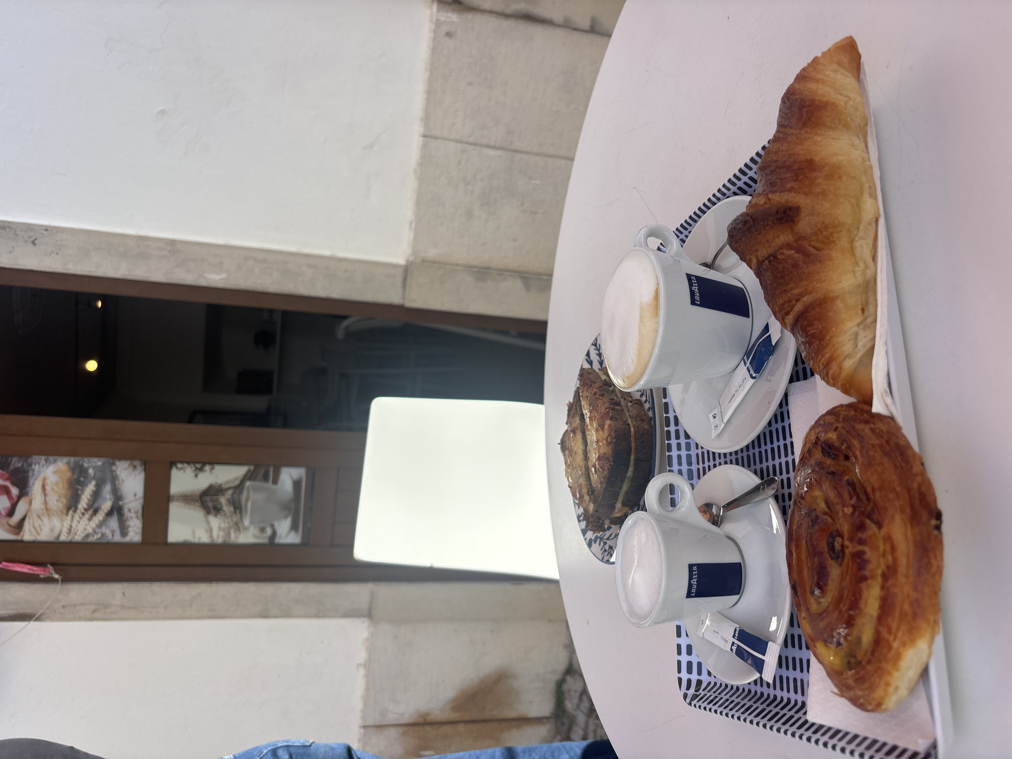 Two cups of coffee with foamed milk and two pastries, one croissant and one spiral-shaped pastry, are arranged on a white tray with a black and white striped border. A rectangular sandwich with melted cheese is on a decorative plate behind the coffee cups. The tray and plate are on a white table. In the background, a storefront with wooden doors and glass panels features images of bread and the Eiffel Tower. A white square board is positioned behind the table. The scene appears to be an outdoor seating area of a cafe in Tavira, Portugal.