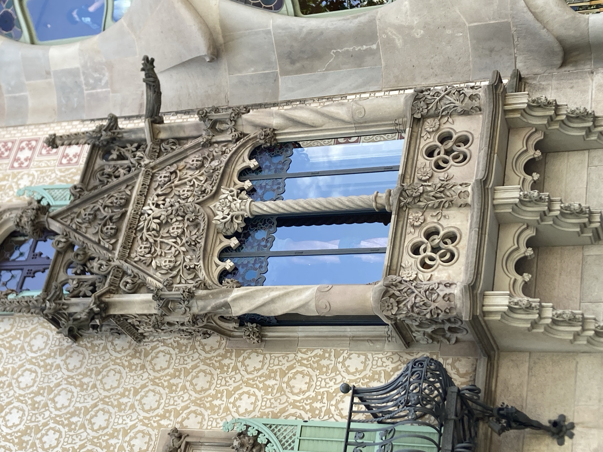 The image displays an ornate, stone balcony of a building in Barcelona, Spain, likely a facade of Casa Batlló given the architectural style. The balcony features intricate carvings of floral motifs and geometric patterns. A twisted stone column supports the structure, and a gargoyle-like spout protrudes from the upper level. The building's wall behind the balcony is adorned with a repeating pattern of stylized circular designs. The window behind the balcony is reflective, showing a clear blue sky. To the left of the balcony, a section of wrought-iron railing and a green-painted window shutter are visible. The stonework of the building is aged, with some visible texture and variations in color.