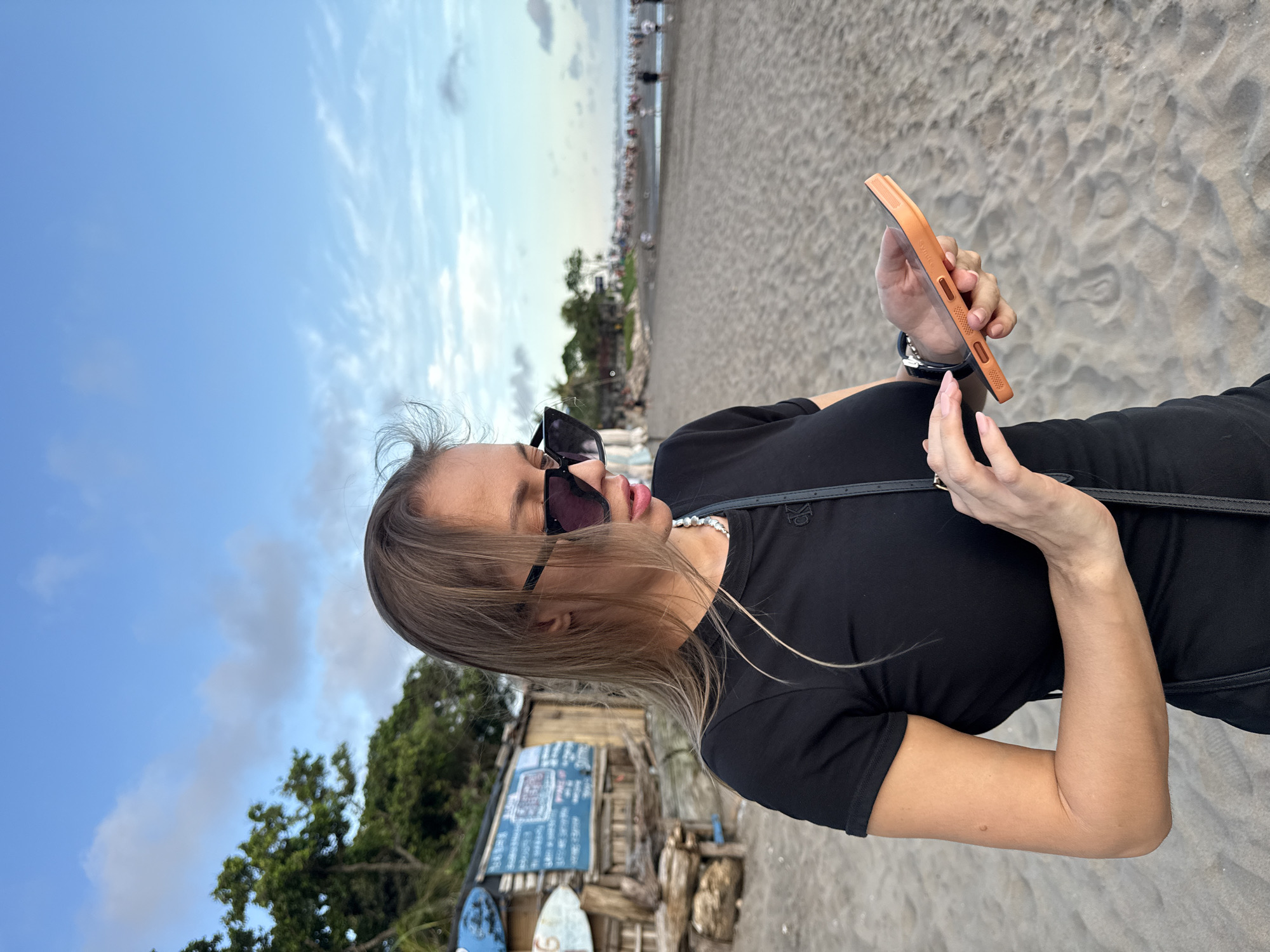 A woman stands on the sandy shore of a beach in North Kuta, Indonesia, facing right and holding a smartphone with an orange case in her hands. She is wearing sunglasses and a black t-shirt with a crossbody purse. Her blond hair is blowing in the wind. In the background, there is a building adorned with surfboards and signs. Further in the background, the beach is crowded with people. The sky is light blue with scattered clouds. The foreground consists of the sandy beach.