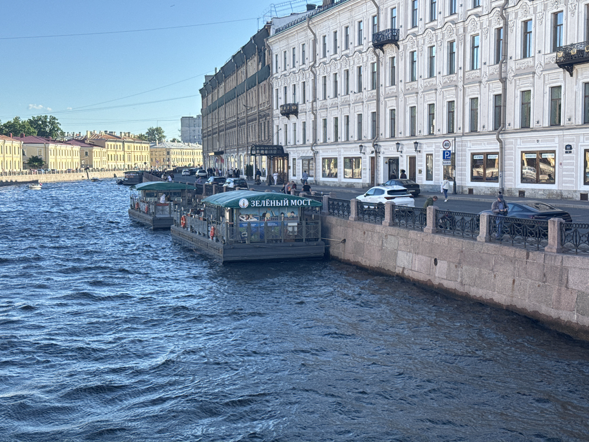This is a scene of the Griboyedov Canal in Saint Petersburg, Russia, on a clear day. A wide, deep blue canal dominates the foreground, with gentle waves rippling across its surface. Moored along the right bank are two tour boats, their awnings providing shade. The first boat, closer to the viewer, is named "ЗЕЛЁНЫЙ МОСТ" (Green Bridge). People are visible on board and on the embankment.
The embankment is lined with a stone wall and a decorative black metal fence. Behind the fence, a sidewalk runs parallel to the canal, populated by several pedestrians and a few parked cars, including a white sedan and a black sports car.
The background features a long, elegant row of classical buildings that characterize Saint Petersburg's architecture. These buildings have multiple stories, numerous windows, ornate facades, and balconies. On the left side of the canal, further down, a section of buildings with yellowish facades and red roofs is visible. The sky above is clear and blue, with no clouds. The overall impression is one of a vibrant urban waterway in a historic European city.
