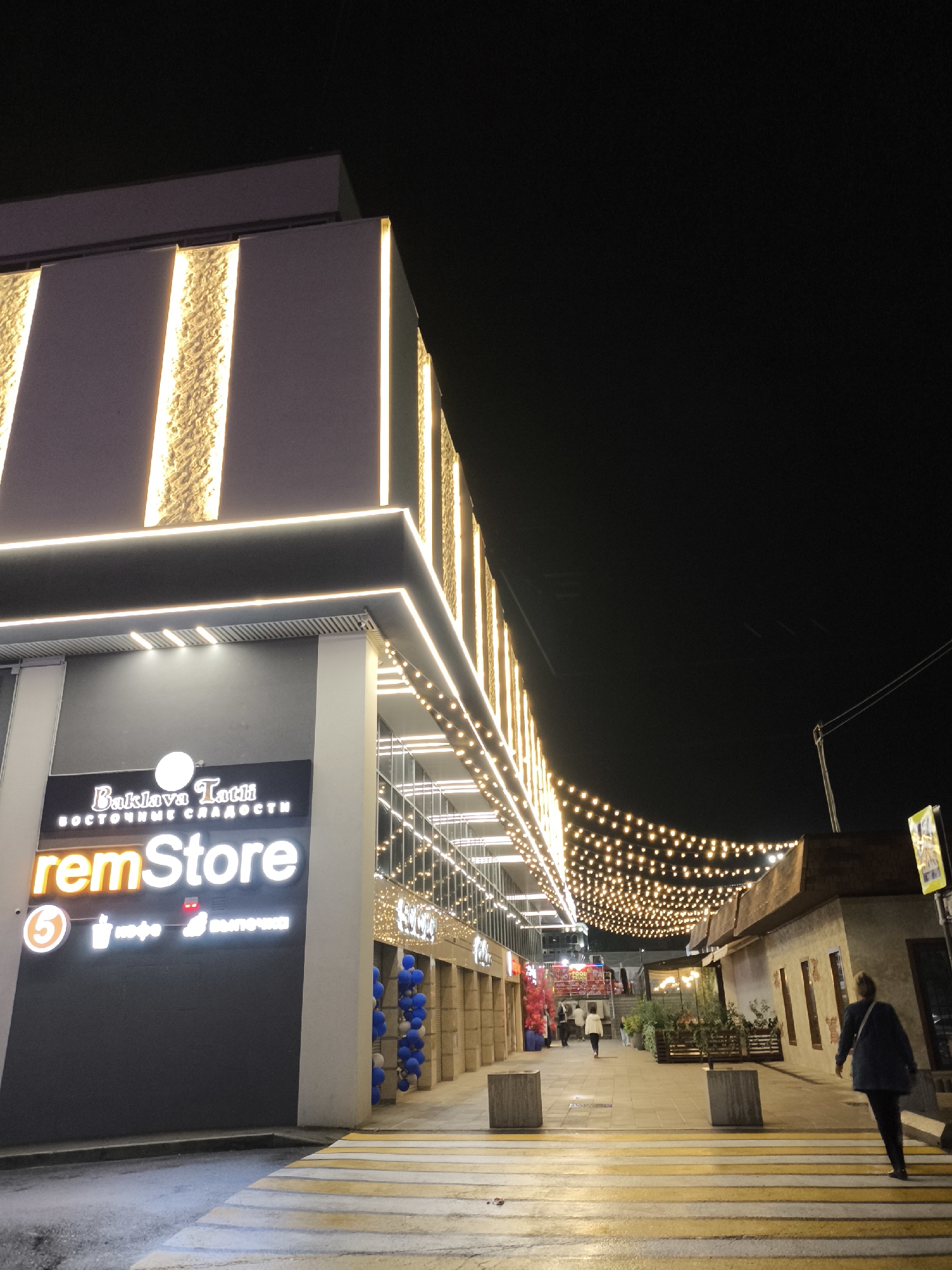 The image depicts an outdoor shopping street at night in Yessentuki, Russia. The scene is illuminated by warm, glowing string lights and architectural lighting. On the left, a modern building facade features vertical, textured panels lit from behind by yellow lights, and a prominent sign reading "remStore" in large white letters, with smaller text above indicating "Baklava Tatli BOCTOЧHЫE CЛAAOCTИ" (Baklava Tatli Oriental Sweets). Below the "remStore" sign are smaller icons and text, one of which appears to be a "5" in a red circle, a coffee cup icon, and an icon of a pastry or bun. Adjacent to this building, a walkway is marked with yellow and white stripes, resembling a pedestrian crossing.
The walkway leads into a pedestrian zone lined with shops and stalls. Another building on the right has a rustic facade with windows and is also decorated with string lights. Several people are visible walking along the street. One person, a woman wearing a dark jacket and carrying a bag over her shoulder, is walking away from the camera across the striped walkway. Other individuals are further down the street, walking towards or away from the shops. Blue and white balloons are clustered near an entrance on the left. The overall atmosphere is lively and festive, suggesting a commercial area popular for evening strolls and shopping. The dark sky overhead is visible, indicating it is nighttime.