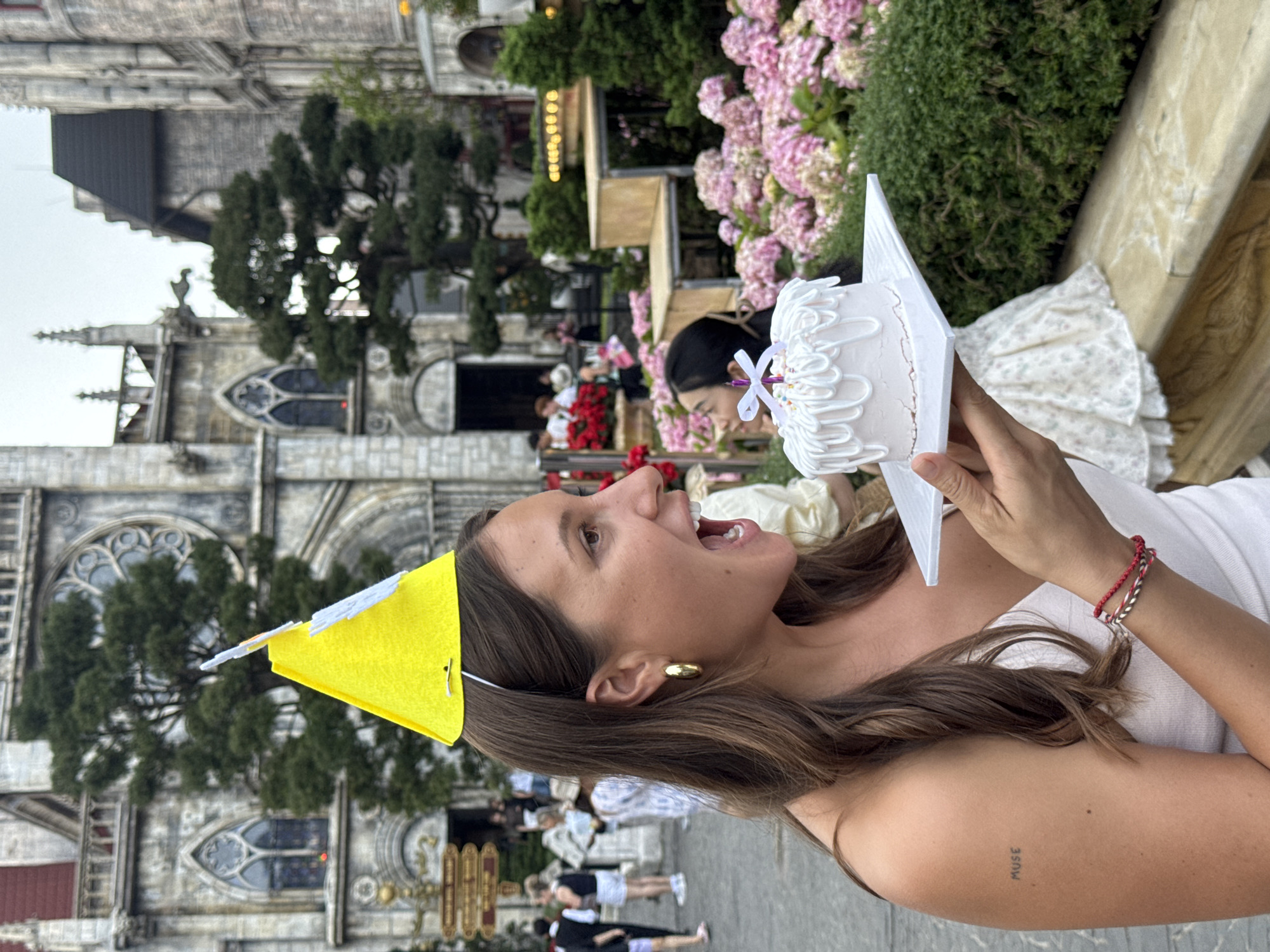 A woman with long brown hair, a yellow party hat, a gold earring, and a tattoo that says "muse" is holding a small white cake with a single lit candle toward her open mouth. She is wearing a white dress and bracelets. In the background, a gothic-style building and a crowd of people are visible, along with flowers and foliage. The location is in Đà Nẵng, Vietnam.