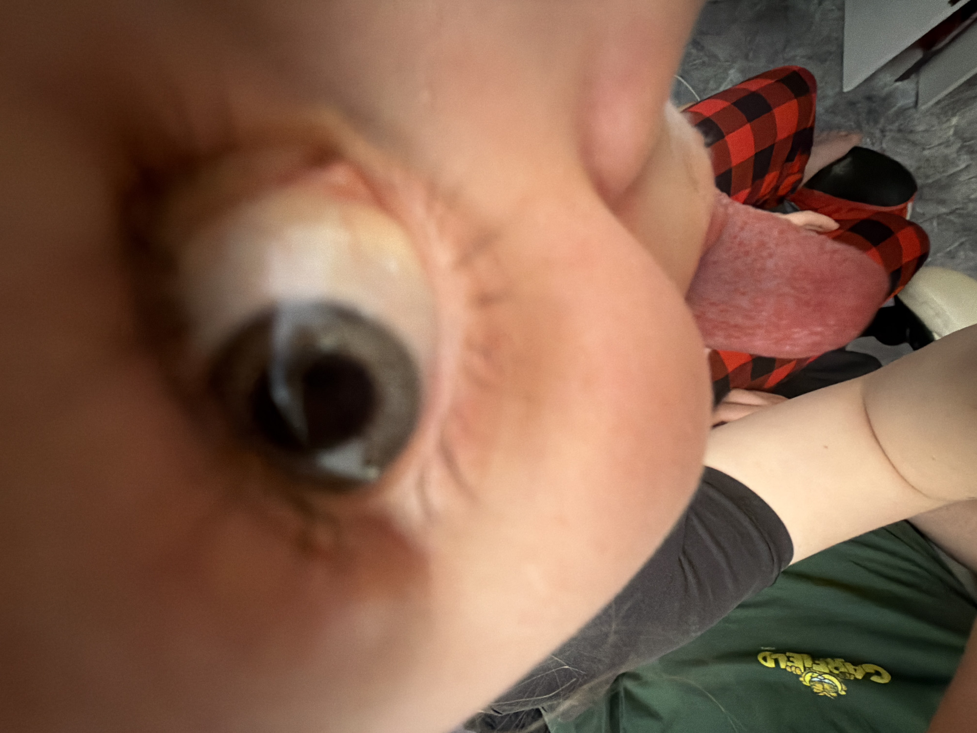 A close-up, distorted view of a person's eye and face dominates the left side of the image. To the right of the face is a partially visible tongue. Further back in the frame, the person appears to be wearing red and black checkered pants. In the lower right corner, part of a green shirt with the word "Garfield" printed on it is visible, along with a portion of what appears to be a black garment or shorts. The background shows a textured grey surface, possibly carpeting, and hints of a white object in the upper right corner. No information is available to determine the location in Perm, Russia.