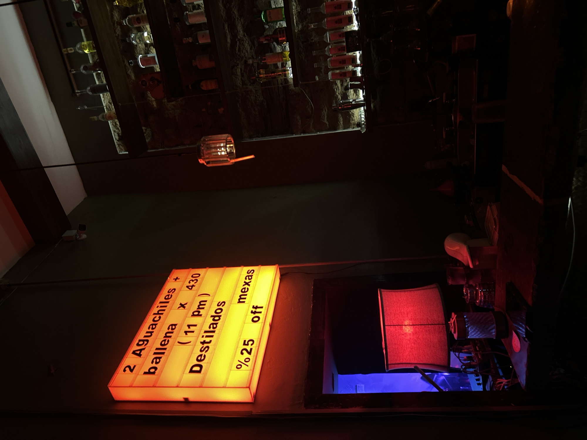 The image captures a scene inside a dimly lit establishment in Mazatlán, Mexico. A sign prominently displays the offer "2 Aguachiles + ballena x 430 (11 pm) Destilados mexas %25 off". The sign is illuminated with a bright yellow light. Behind the sign is an opening/window that leads to a darker space featuring a red lamp. The overall ambiance suggests a bar or restaurant, possibly after dark. Above, on the ceiling, are multiple shelves holding rows of bottles. The lighting is predominantly artificial and creates a stark contrast between the bright sign and the shadowy corners of the interior.