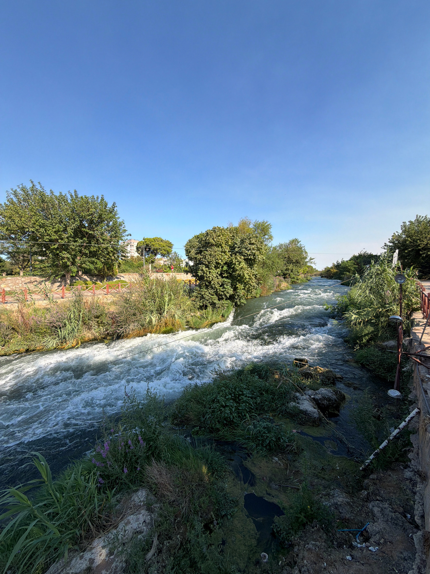 The image depicts a flowing river with white rapids, bordered by lush green vegetation and trees. In the background, buildings are visible, suggesting an urban or semi-urban setting. A red wooden railing runs along the right bank of the river, with a pathway visible on the left bank. The sky is a clear blue. This scene is located at the Düden Waterfalls in Antalya, Turkey.