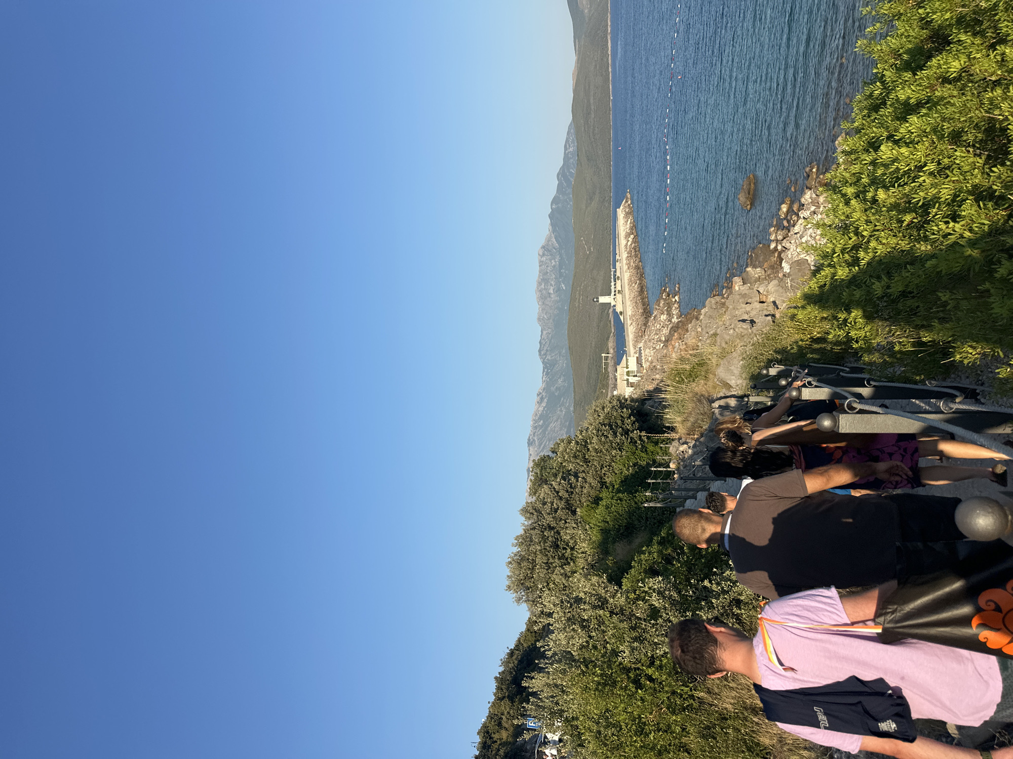 A group of people walks along a coastal path with lush green vegetation on the left and the blue sea on the right. In the distance, mountains form a backdrop under a clear blue sky. The path is bordered by a metal railing and is made of stone. Some of the people are carrying bags. A lighthouse and a breakwater are visible in the distance along the coast. The scene appears to be in a sunny, outdoor environment, possibly a tourist area. The location is identified as Radovići, Montenegro.
