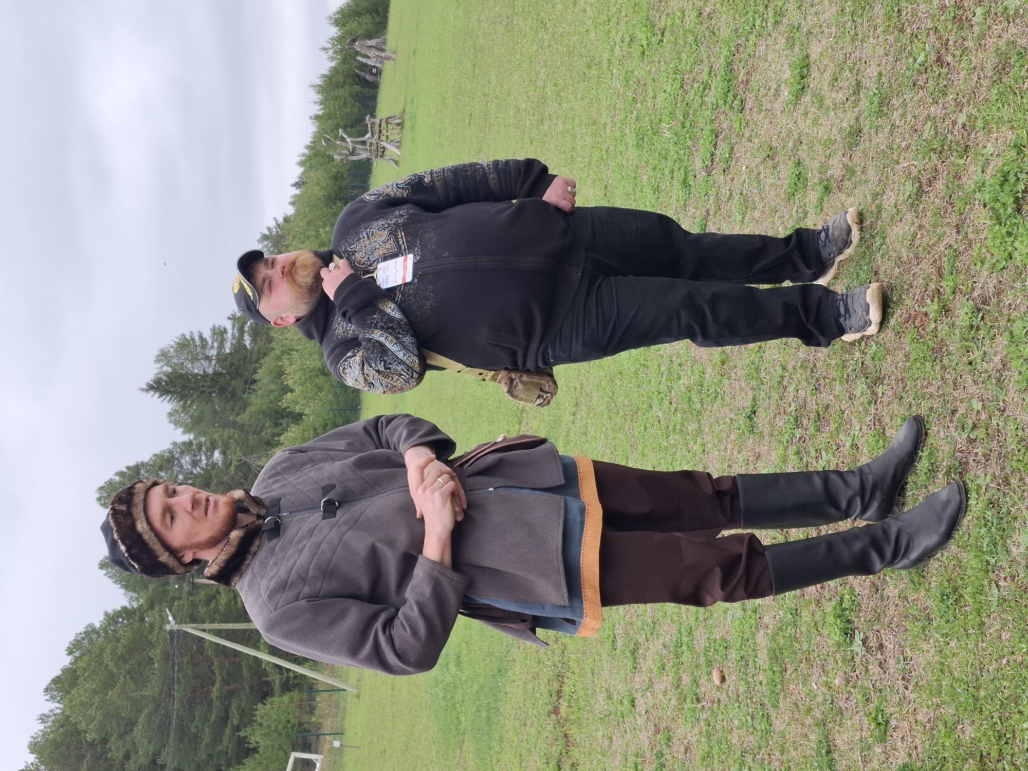 Two men are in a grassy field. One man is wearing a fur hat, brown pants, and black boots. He is wearing a grey coat. The other man is wearing a black cap and black pants and a black jacket. He is standing behind the first man. The weather is overcast. In the background, there are trees.