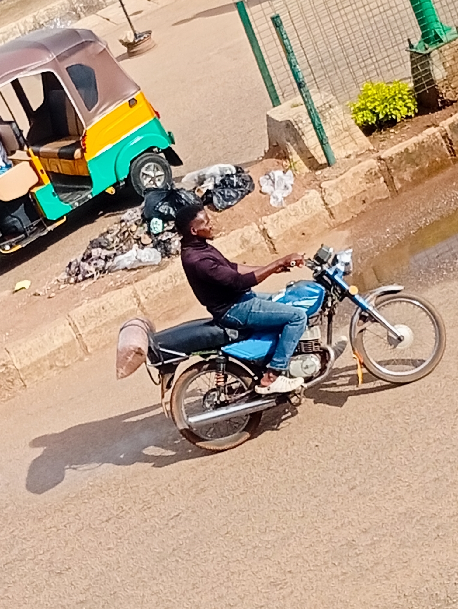 In Bukuru, Nigeria, a man rides a blue motorcycle on a dirt road. He is wearing a maroon turtleneck shirt and blue jeans. He is wearing white slip-on shoes. The motorcycle has a brown sack strapped to the back. To the left of the motorcycle is a multicolored auto-rickshaw, which is green, yellow, and brown. There is trash and debris on the ground near the auto-rickshaw. Behind the motorcycle and auto-rickshaw is a concrete curb and a dirt embankment. A fence and a small green bush are visible on top of the embankment. The sky is visible in the upper left corner of the image.