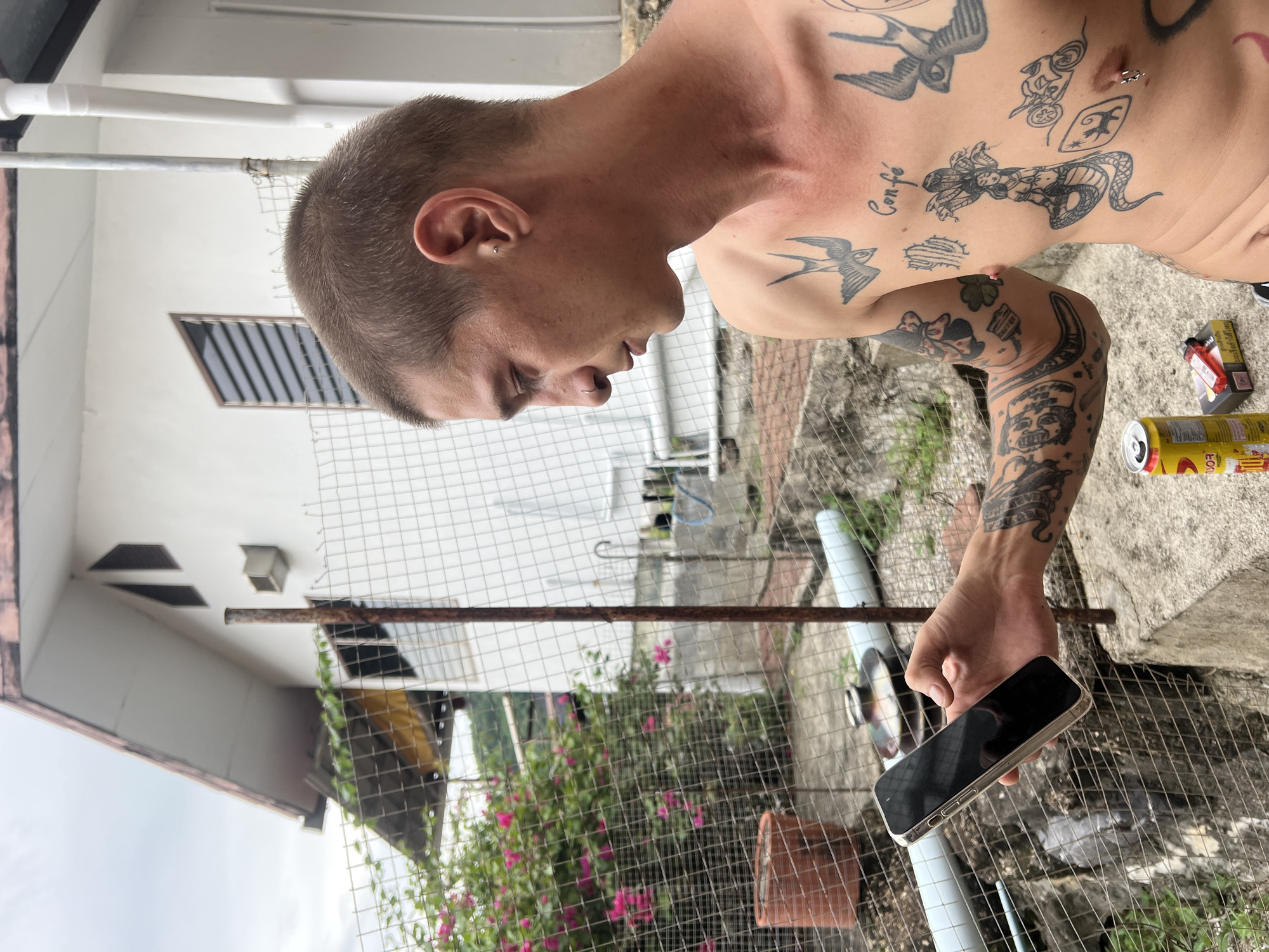 A man with tattoos on his chest and arms is holding a smartphone in his right hand. He is standing outdoors, with a white house and a chain-link fence in the background. There are pink flowers and a terracotta pot visible behind the fence. A yellow can and a pack of cigarettes are on the ground in front of him. The man is looking down at the phone, and appears to be in profile.