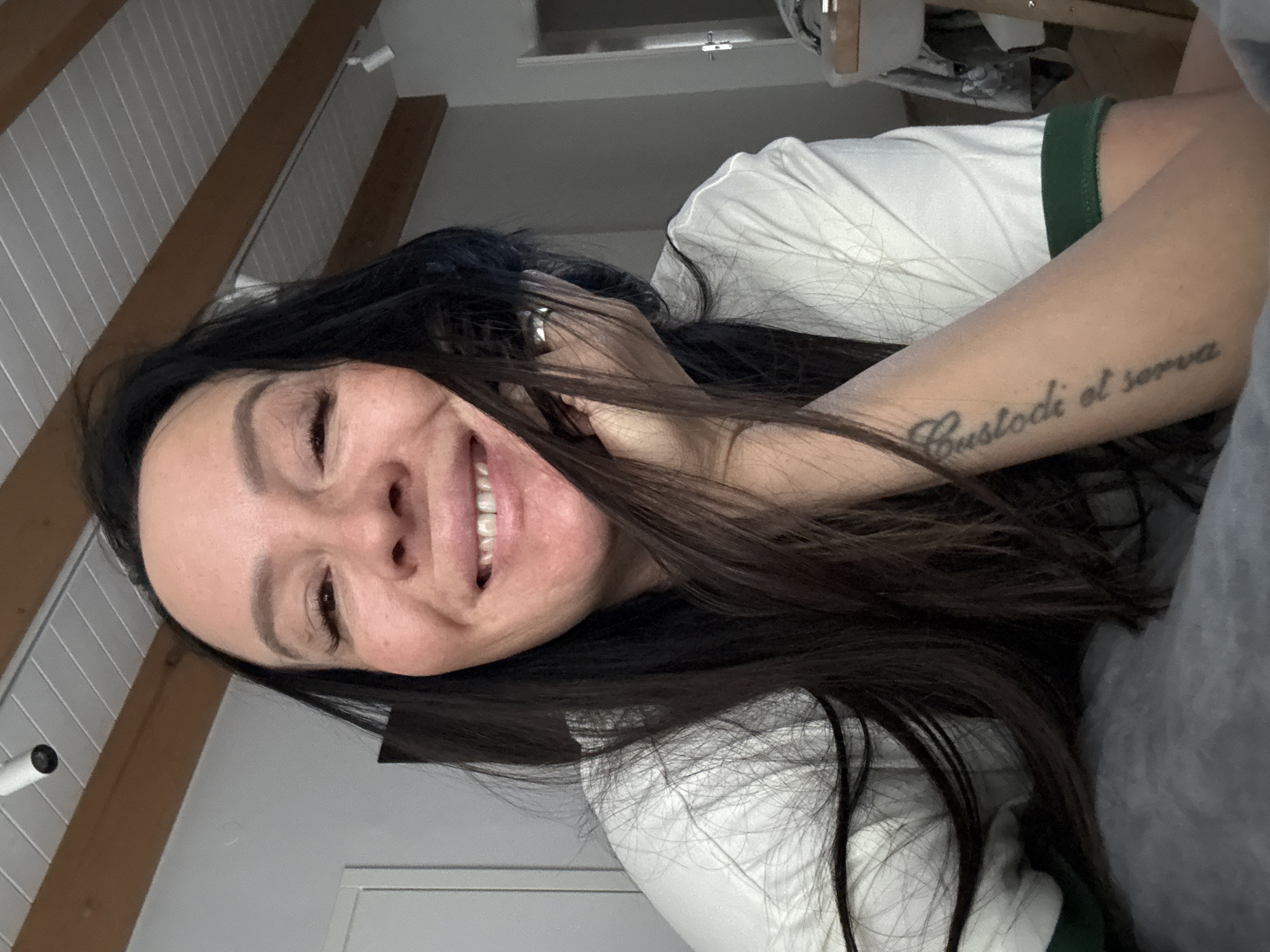 A woman with dark hair is smiling and looking at the camera. She is wearing a white t-shirt with a green collar. There is a tattoo on her arm that reads "Custodi et serva". The background is blurry, but it appears to be a room with white walls and wooden beams. There is no visible information to suggest that the location of the image is the city of Chaikovsky, Russia.