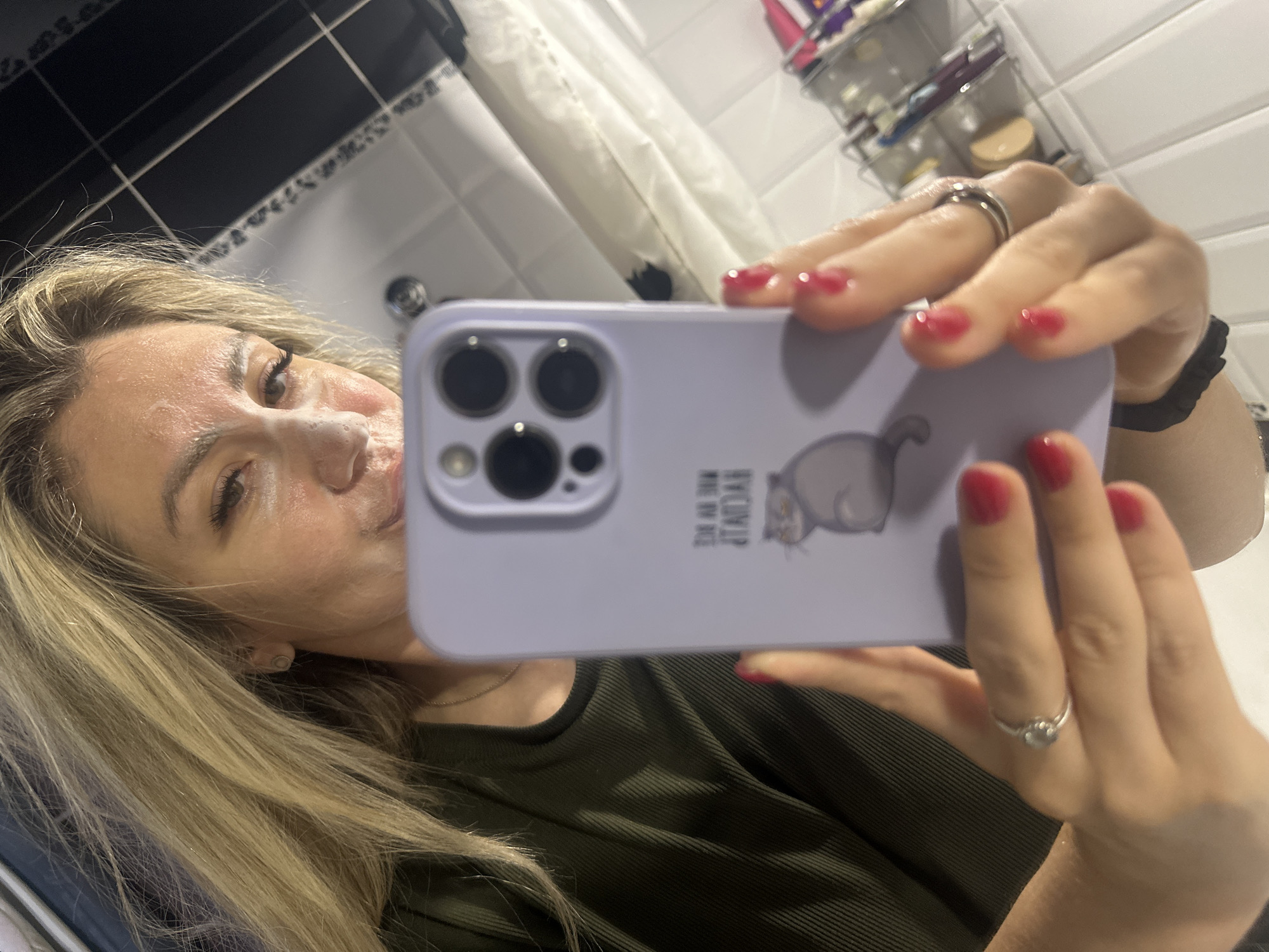 A woman is taking a selfie in a bathroom. She has blonde hair and is wearing a green top. Her face has a white face mask on it. She is holding a light purple phone in her right hand, which has a picture of a cat on the back. Her fingernails are painted red. She is wearing two rings on her left ring finger. The background of the bathroom shows white tiles and a shower curtain. The image was taken in the city of Novoaltaysk, Russia.