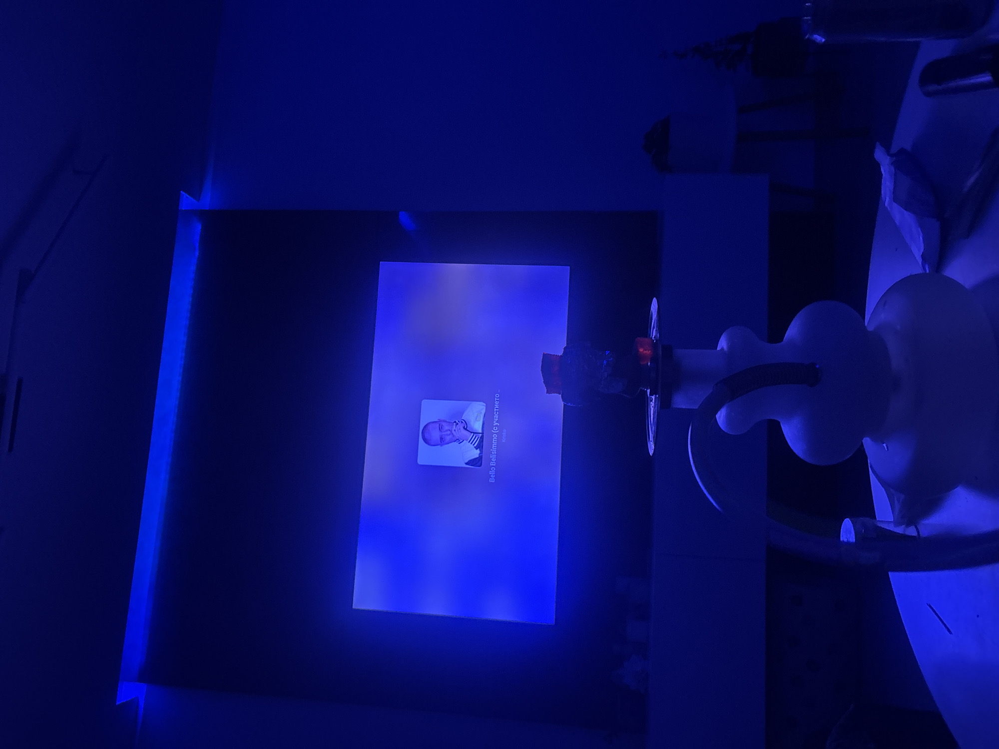 In an dimly lit room with pervasive blue lighting, a hookah with a white base and black hose sits on a table. A television screen displays a profile picture of a man with the name "Bella Bellissimo" and "Krsika" below it. The screen is illuminated by blue light, which also reflects off the walls and furniture. The overall scene suggests a relaxed, indoor environment.