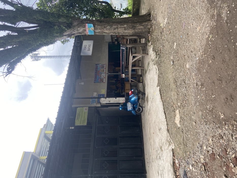 A blue scooter is parked in front of a building with a sign that says "TOKO BATAS" above a shop window, a tree with a sign that says "TOKO BATAS" and a second sign with indecipherable writing is on the right side of the building. The building is in a state of disrepair, with a large crack in the concrete. There is a wooden table and bench in front of the building. The scene is in Ciparay, Indonesia, at the coordinates -7.022 latitude, 107.68 longitude. There are no people present.
