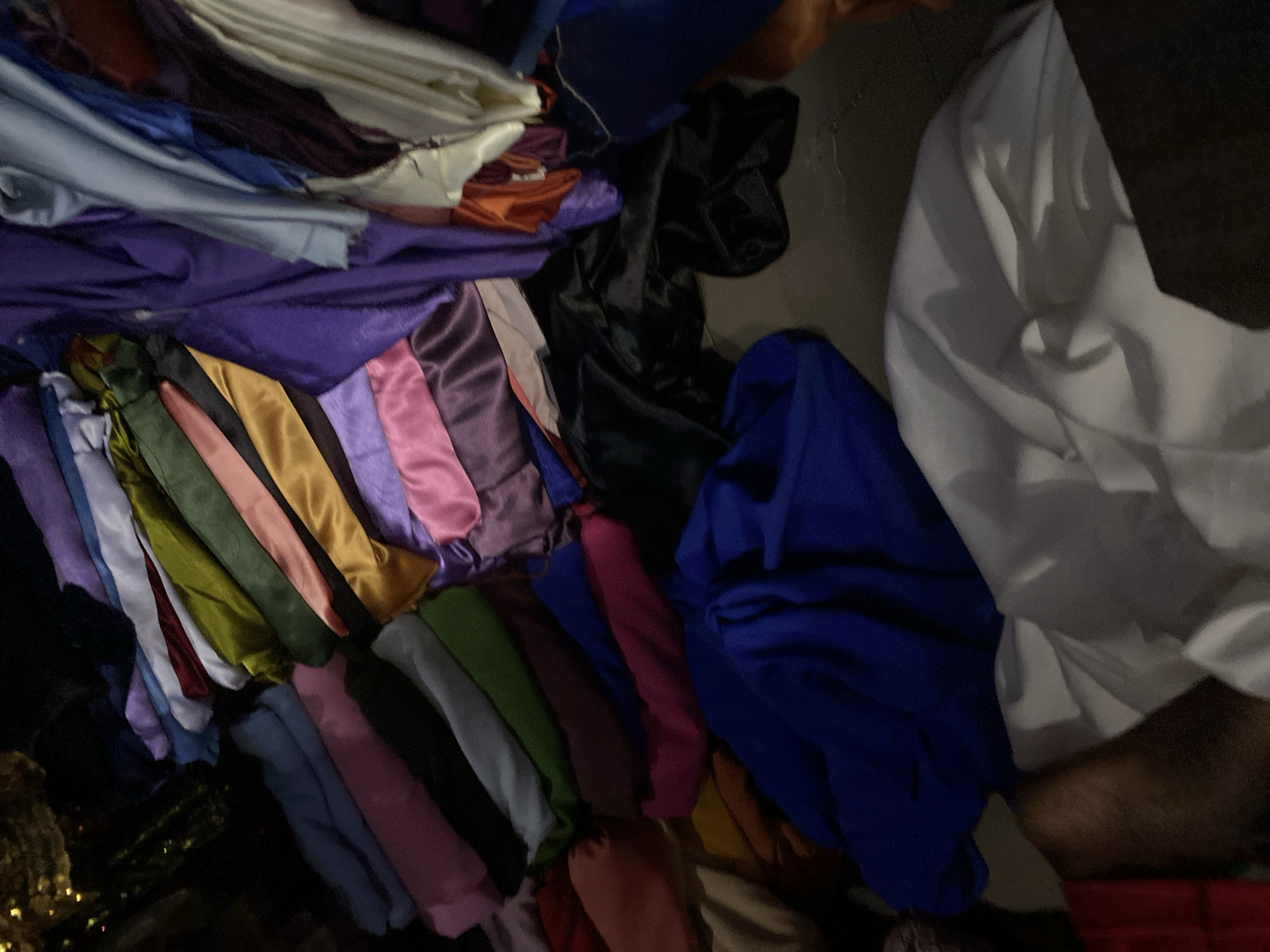 The image depicts a pile of colorful fabrics, likely silks or satins, with various shades of purple, pink, green, blue, yellow, and white visible. The fabrics are draped and folded, creating a textured and somewhat chaotic arrangement. Some of the fabrics are smooth and shiny, while others appear more matte. The lighting is dim, casting shadows and highlighting the sheen of some materials. The scene is likely indoors, possibly in a market or storage area, given the quantity and variety of textiles. No specific landmarks or context are provided to pinpoint a precise location within Jalingo, Nigeria, beyond the general setting of stored textiles.