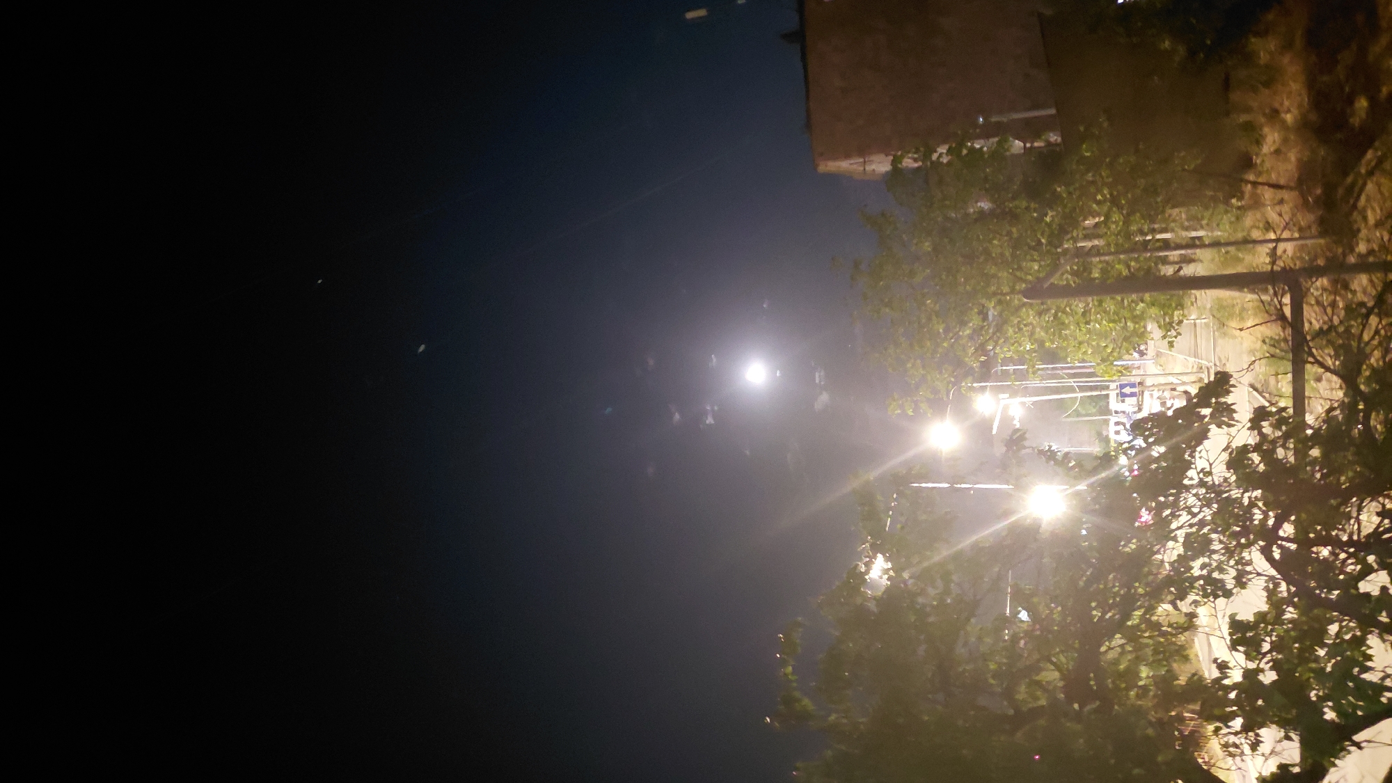 The image depicts a nighttime scene on a street in Abovyan, Armenia. The sky is dark with a bright moon visible, partially obscured by some clouds or atmospheric haze. Several streetlights illuminate the scene, casting bright beams that cause lens flare. Trees with green foliage are in the foreground, with some branches extending into the frame. In the background, there are buildings, including one with a brick facade on the right side of the image. There are also some vehicles and what appear to be people on the street in the distance. The overall impression is of a moderately lit urban street at night.