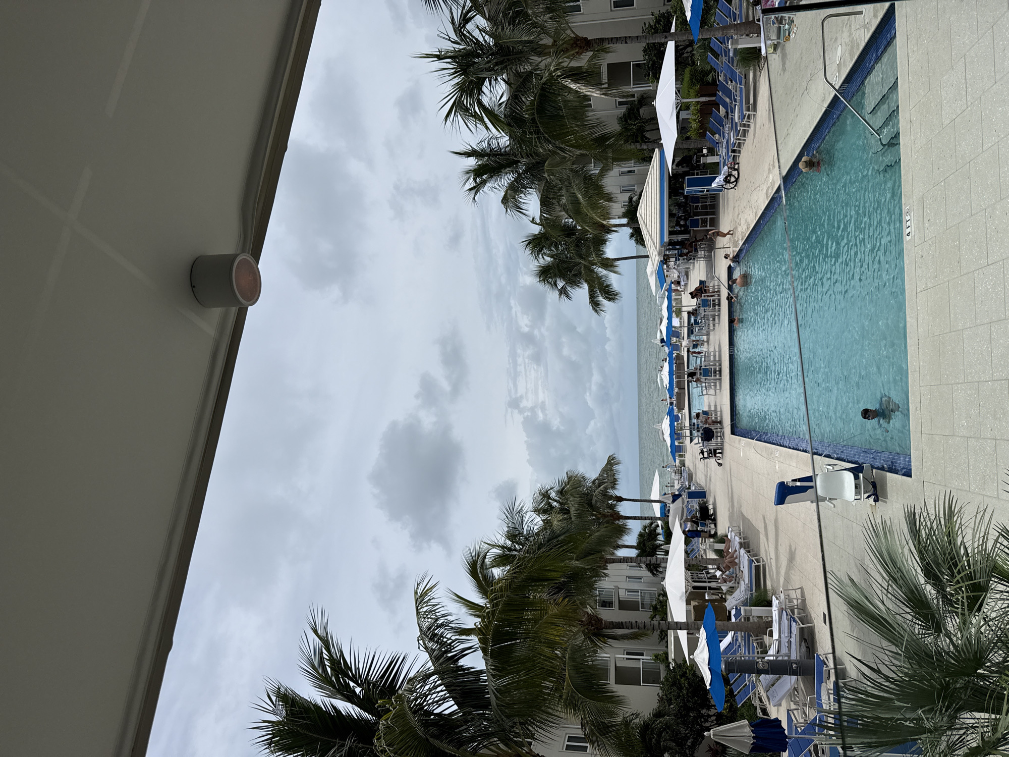 This is a photograph of a resort pool area, likely in Naples, Florida, United States of America, given the tropical vegetation. The image is taken from an elevated, covered vantage point, looking out over the pool and towards the ocean.
In the foreground, a large, rectangular swimming pool dominates the scene. The water is a vibrant turquoise. Several people are visible in and around the pool. One person is swimming in the deeper section of the pool, while others are in the shallower end, with one person appearing to be a child playing in the water. A pool lift mechanism is visible on the edge of the pool.
Surrounding the pool are paved deck areas with lounge chairs and umbrellas. Many lounge chairs are occupied by people relaxing. Buildings with balconies and windows line the sides of the resort area, and numerous palm trees are scattered throughout, indicating a warm climate. A white canopy structure with blue trim provides shade in the background, and it appears to be a bar or food service area.
In the distance, the ocean is visible under a cloudy sky. The overall atmosphere is one of a relaxed resort vacation. The sky is overcast with scattered clouds, suggesting the possibility of rain or a break in the sun. The lighting is diffused, typical of an overcast day.