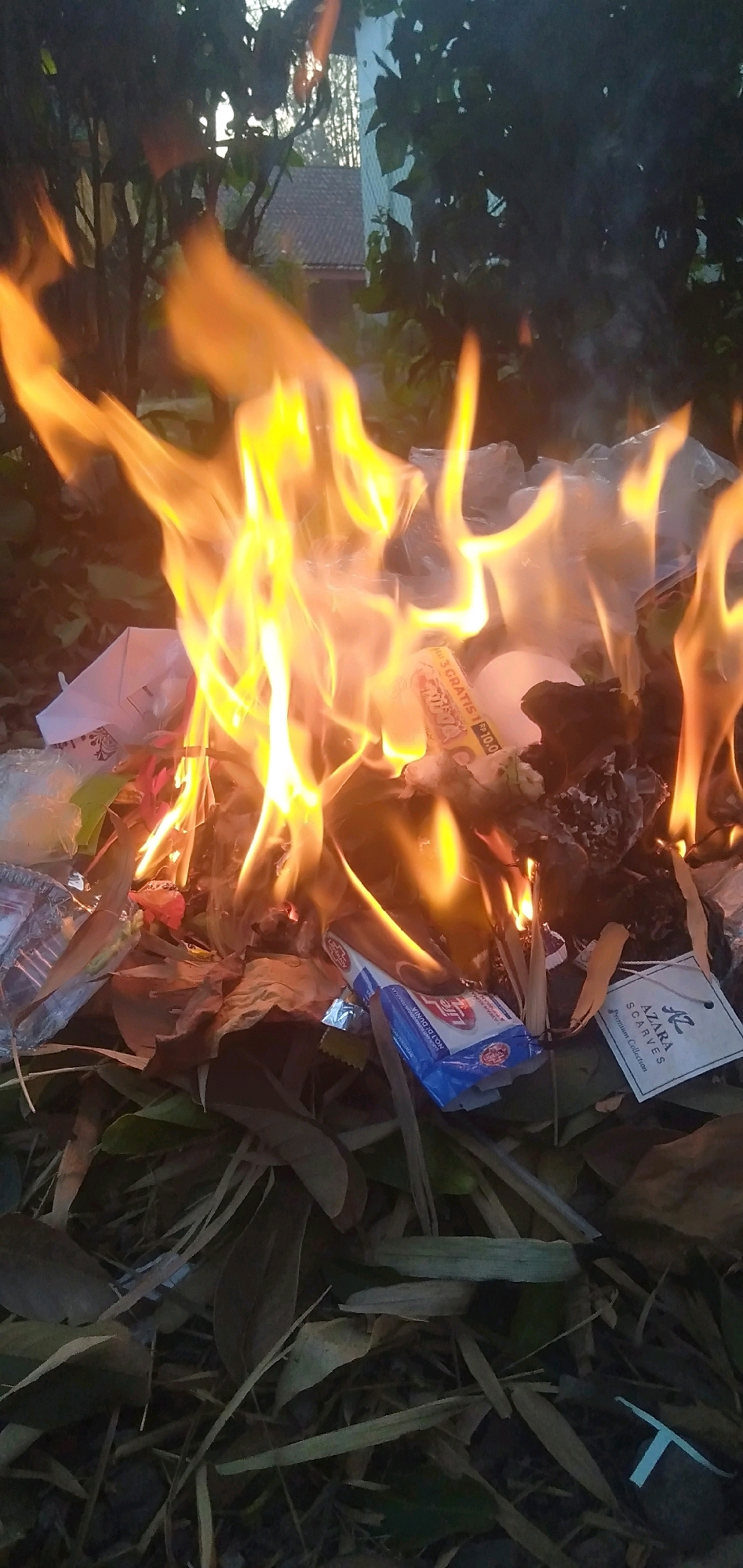 In Plemahan, Indonesia, a fire burns among dry leaves and assorted debris. The flames, a vibrant orange and yellow, are the focal point of the image, consuming what appears to be discarded items. Among the burning materials, a white tag with "AZARA SCARVES Premium Collection" printed on it is visible. Also present is a blue and white package, possibly a product wrapper, with text that is partially legible. The background is obscured by dark foliage and the indistinct outline of a building with a tiled roof. The overall impression is one of an outdoor setting where refuse is being incinerated.