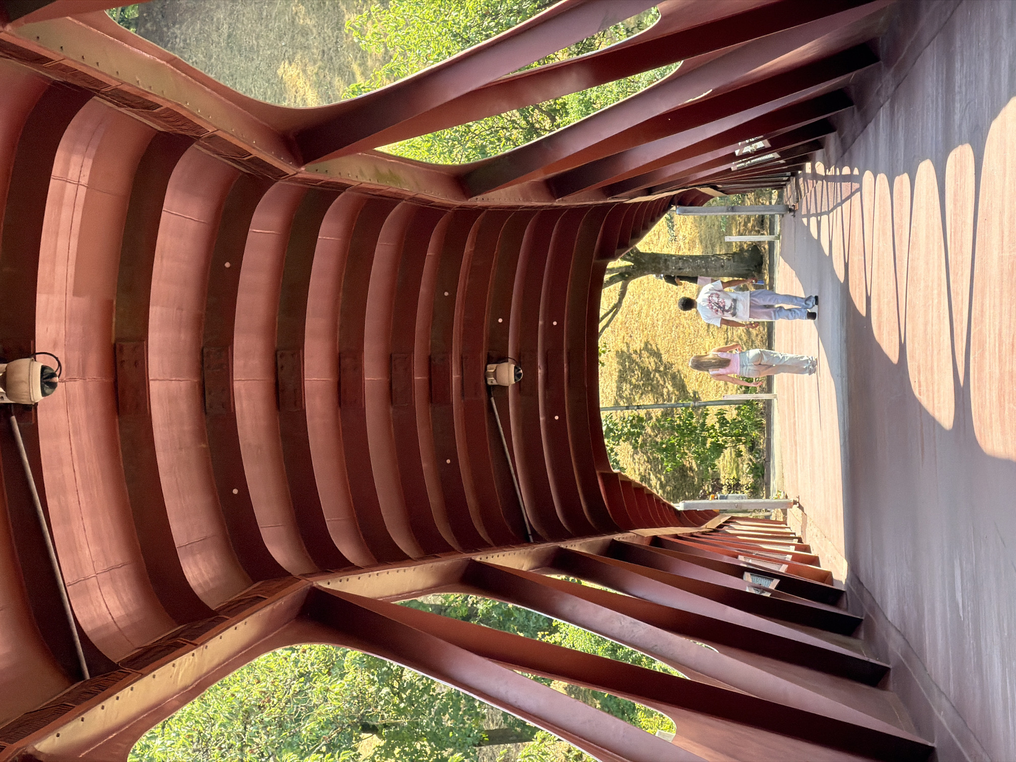 Two individuals, a male and a female, walk away from the camera on a modern red pedestrian bridge. The bridge has a distinctive curved, ribbed ceiling structure made of dark red metal beams. The sunlight casts long, heart-shaped shadows on the bridge's surface. To the left and right of the bridge, lush green trees and foliage are visible. The background shows a gentle slope covered in dry grass, typical of the landscape surrounding the "Pont du Moulin" in Esch-sur-Alzette, Luxembourg, a location known for its industrial heritage and revitalization efforts. Security cameras are mounted on the underside of the bridge's ceiling. The atmosphere is serene and bathed in natural light.