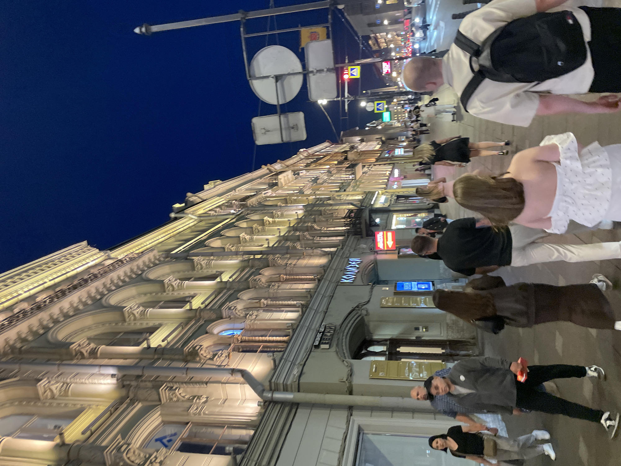 This detailed image depicts an evening urban street scene on Nevsky Prospekt in Saint Petersburg, Russia. The sky is a clear, dark indigo blue.
On the left, a multi-story historical building with a light-colored, ornate facade dominates the view. Its classical architecture features numerous arched windows, decorative columns, pilasters, and cornices, all brightly illuminated by upward-angled lights. A vertical grey downspout is affixed to the building's exterior. Signs on the ground floor display Cyrillic text, including "БИЗНЕС ЦЕНТР ЛЕВСКИЙ" (Business Center Levsky) and "КОЛИЗЕЙ" (Kolizey).
The wide sidewalk is densely populated with pedestrians. In the foreground, on the right, a man wearing a beige shirt and a black backpack walks away from the camera. Next to him, a woman in a white off-the-shoulder top and white skirt also walks away. Slightly to their left and closer to the camera, a man in a dark grey zipped jacket and black pants, holding a small red object, walks towards the camera. Behind him, a woman in a black t-shirt and light grey pants, carrying a light brown shoulder bag, walks towards the camera. Further into the mid-ground, a woman with long dark hair, wearing a dark long-sleeved top and a dark long skirt, walks away from the camera. Numerous other indistinct figures are visible walking on the sidewalk further down the street.
Above the street on the right, a series of traffic signs are suspended from a metal framework. These include a large circular sign (seen from the rear), a rectangular sign depicting a deer or elk, and a smaller rectangular sign with yellow and red elements. Traffic lights are visible, with one displaying a red light for vehicles and another showing a white pedestrian crossing symbol. Vehicles with illuminated headlights and taillights are visible in the distance on the street. A tall, slender pole with a pointed top stands against the dark sky on the far right. The scene is illuminated by the building's facade lighting, streetlights, and various commercial signs, casting a warm glow on the street level.