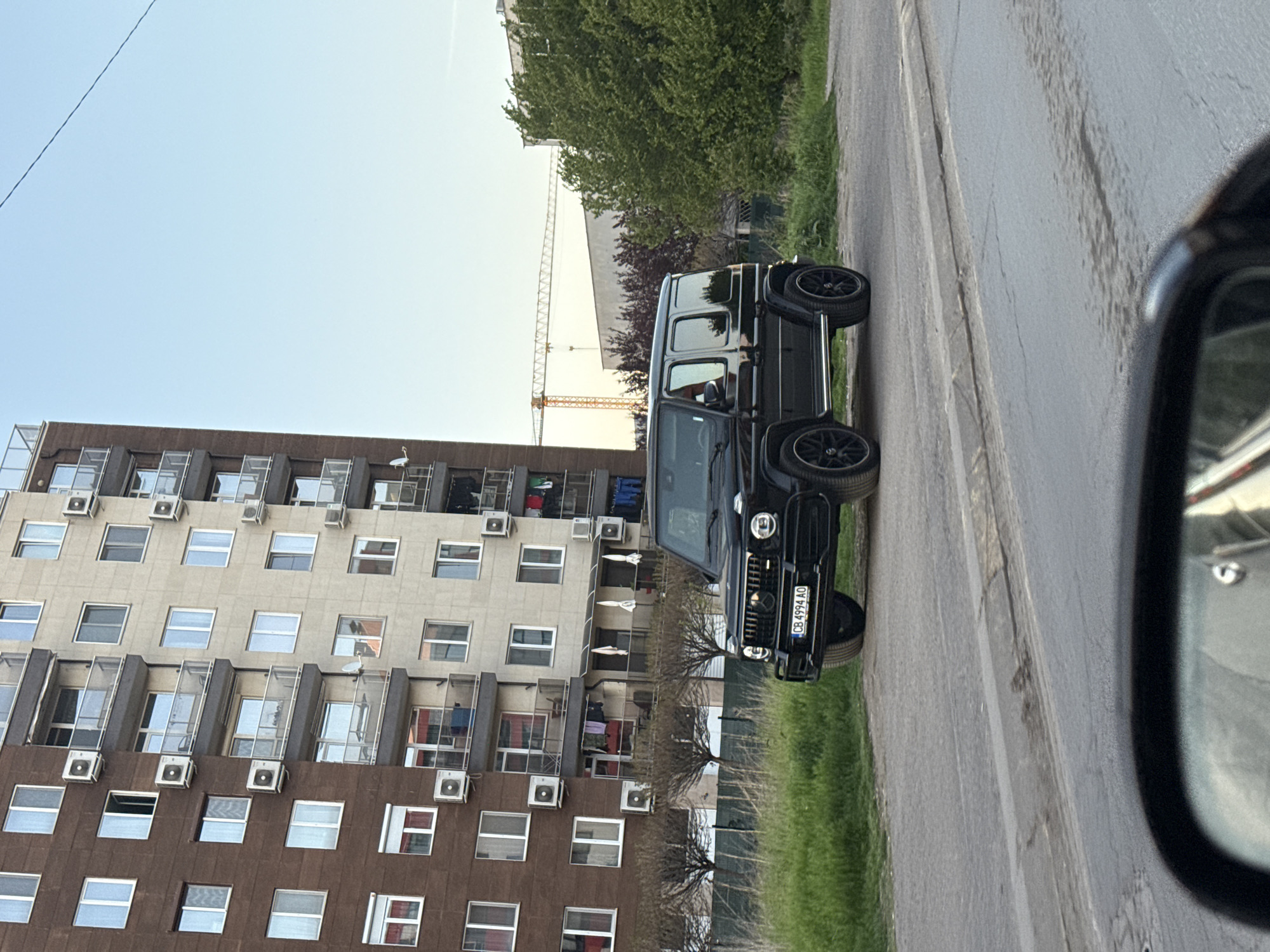 The image shows a black Mercedes-Benz G-Class SUV parked on a street next to a multi-story residential building. The building has many windows and air conditioning units. There is a construction crane visible in the background. Part of a car's side mirror is visible in the right foreground. The SUV's license plate reads "CB 4994 AO," indicating it is registered in Bulgaria. The location is likely in Sofia, Bulgaria, based on the license plate and the style of the residential building. The street is paved, and there is some grass and vegetation adjacent to it. There are no people visible in the frame.