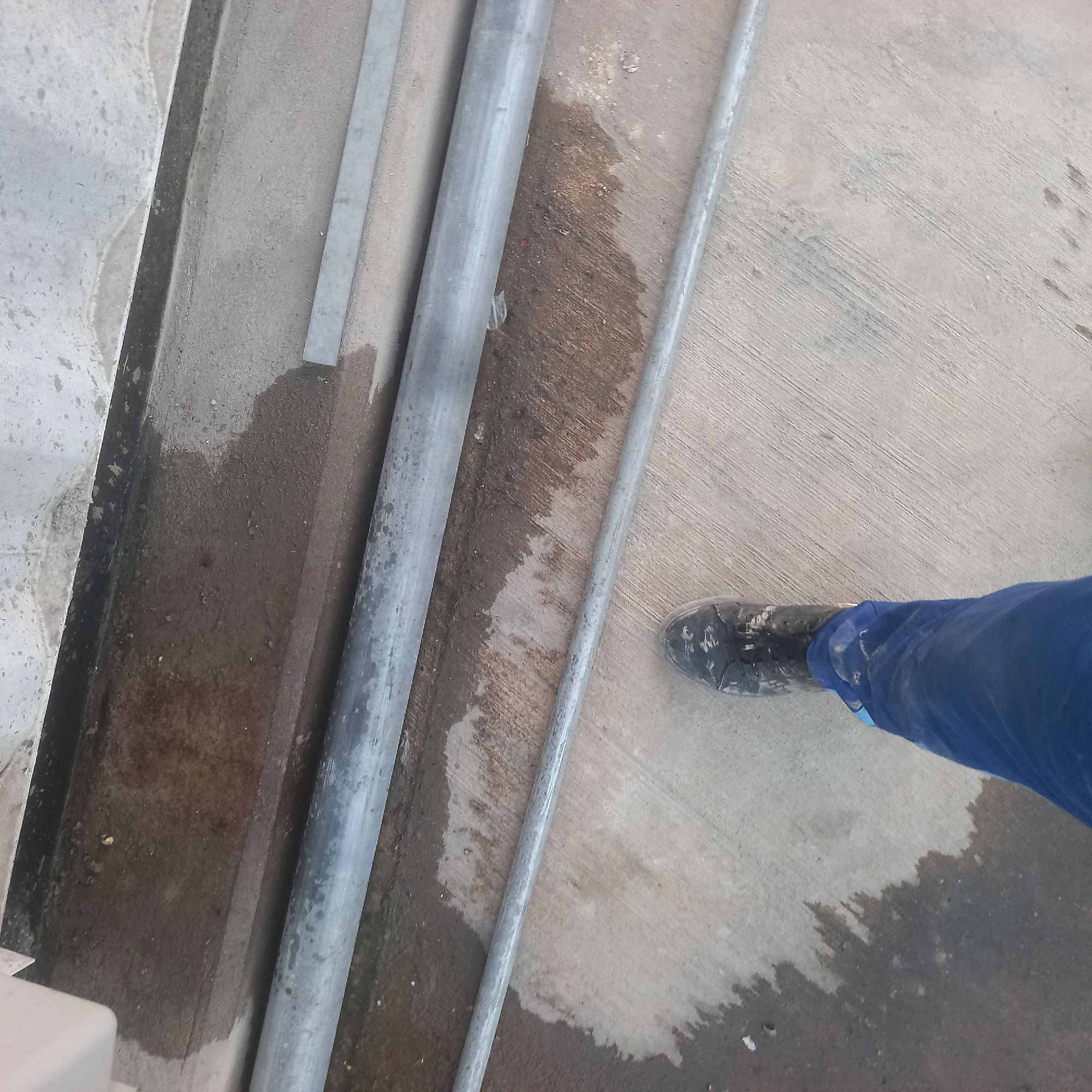 The image depicts a section of a concrete floor with several lengths of metal piping laid horizontally. A person, wearing dark-colored work boots and blue work pants, is standing in the lower left portion of the frame, with their left leg and foot visible. The concrete surface appears somewhat wet or stained in patches, suggesting recent moisture. In the background, a dark, recessed area runs along the top edge of the frame, and above that, a lighter concrete surface. This scene is consistent with an outdoor industrial or construction setting, possibly in Bububu, Tanzania, given the context of the query.