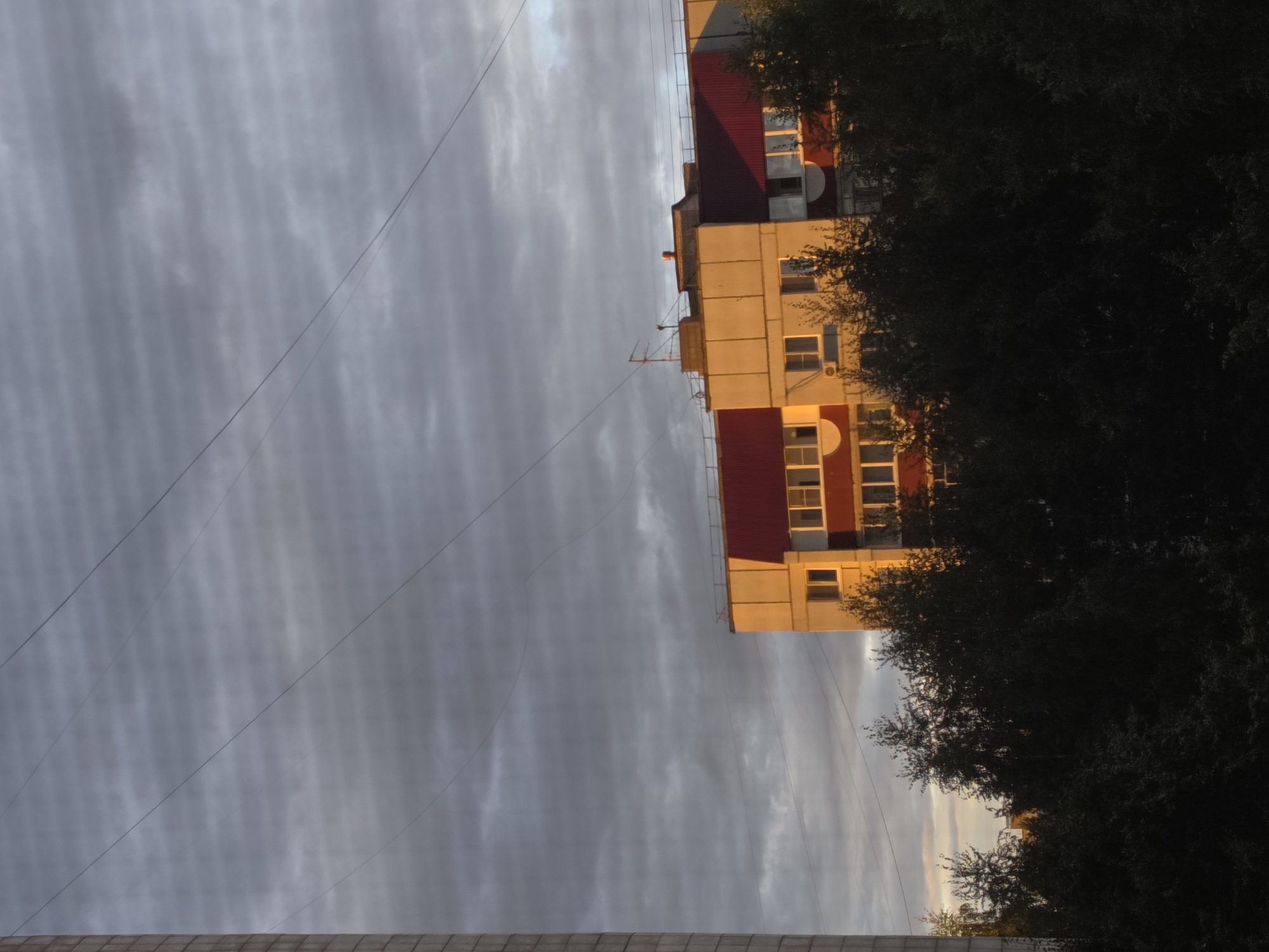 A concrete building, likely a residential apartment block, is visible on the left side of the image. A multi-story building with a red roof and light-colored facade is situated to the right, illuminated by the golden light of sunset. The building has multiple windows and visible architectural details, including rounded elements under some windows. A dense line of dark green trees occupies the foreground, obscuring the lower portions of the buildings. The sky is filled with cloudy, striated patterns, suggesting a partially overcast evening sky, with thin power lines crisscrossing the frame. The scene appears to be an urban residential area in Chaikovsky, Russia, during twilight.
