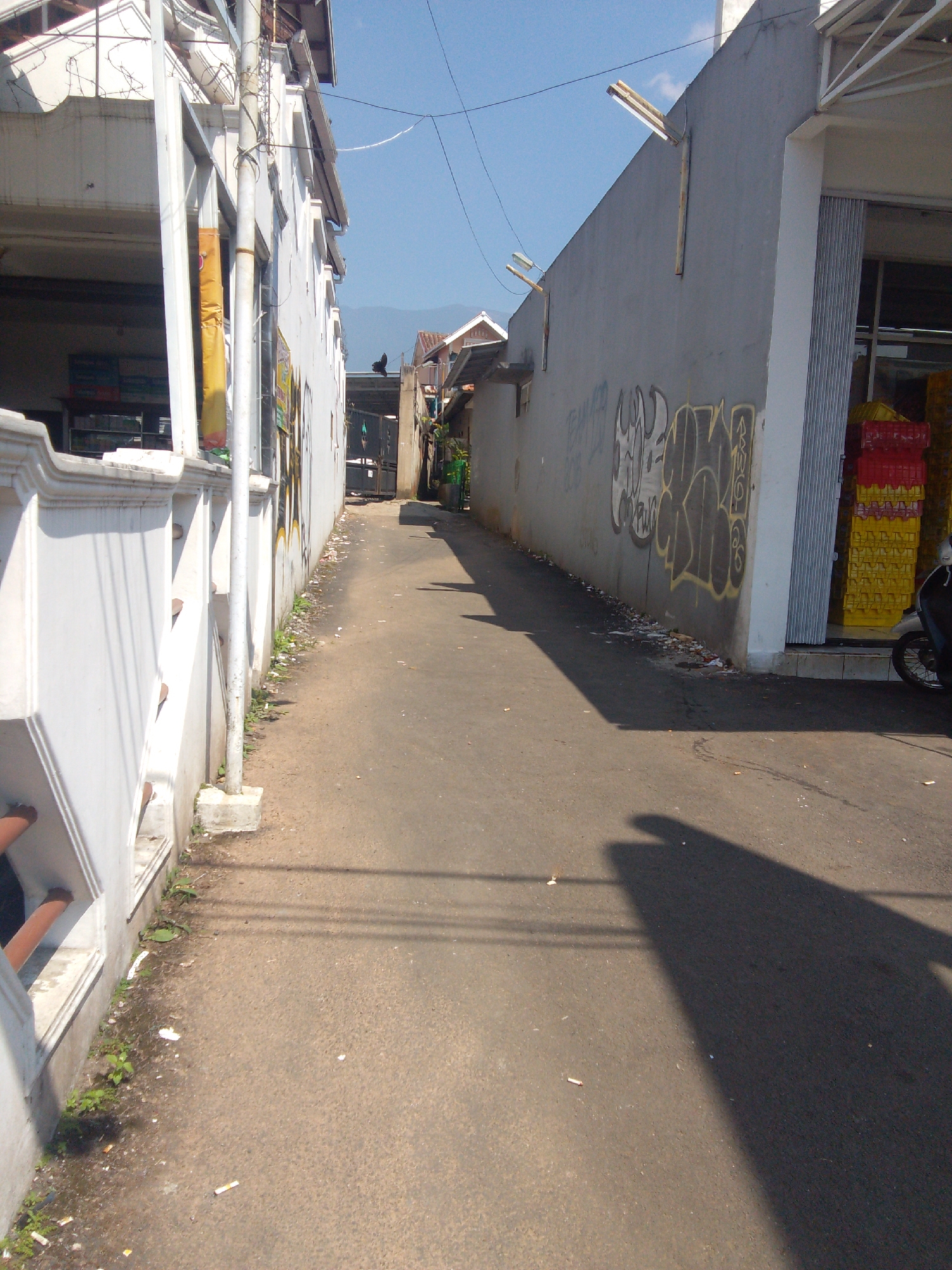 This is a street-level photograph taken outdoors on a sunny day, likely in a residential or commercial area of Cianjur, Indonesia. The perspective is from the ground, looking down a narrow asphalt road flanked by buildings on both sides. On the left, a white building with a decorative railing extends along the foreground. To the right, a gray building with graffiti on its wall runs parallel to the road. The buildings appear to be two to three stories high. The road shows signs of wear and tear, with some debris and cigarette butts scattered on the surface. In the distance, the road curves slightly, leading towards a gate or entrance. Beyond the buildings, a clear blue sky is visible with some faint electrical wires crossing overhead. A single motorcycle is parked on the right side of the road, near the entrance of the gray building. The overall scene depicts an ordinary urban environment, possibly a back alley or a less-trafficked street.