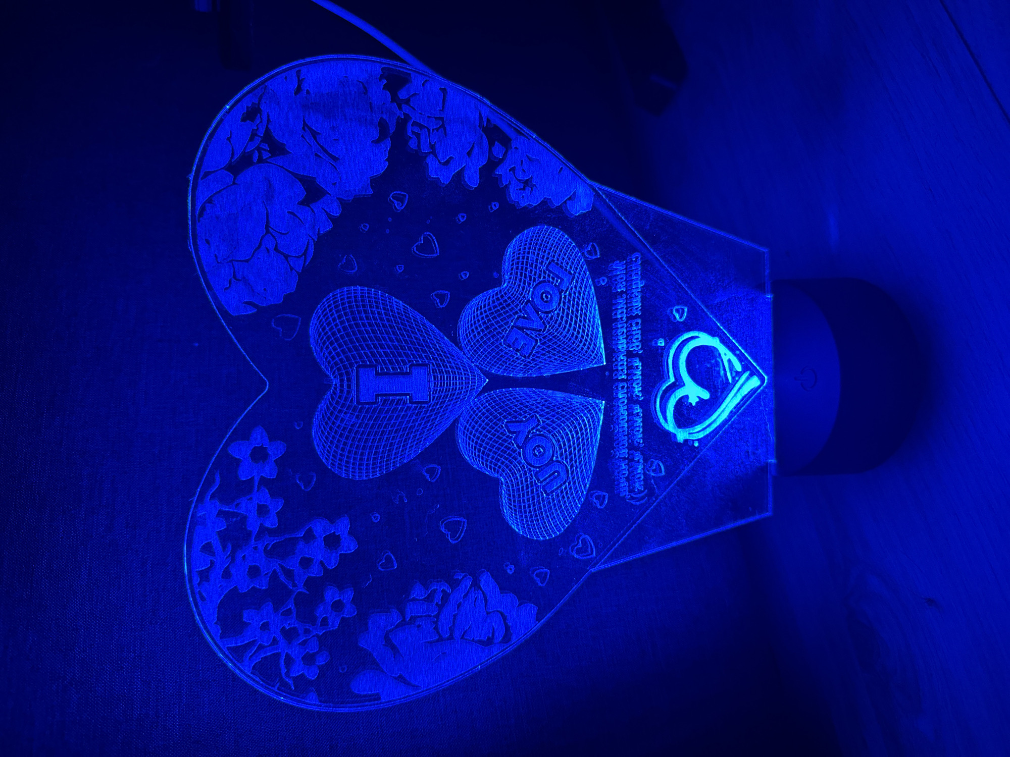 This is a media file of a heart-shaped acrylic lamp illuminated with blue light, resting on a dark, textured surface, possibly fabric, and a wooden surface to the right. The lamp itself is intricately designed, featuring etched floral patterns along the edges of the heart and three smaller hearts within the larger shape. Two of the smaller hearts are etched with the word "LOVE," and one has the letter "I" etched in it. Another smaller heart etched with "JOY" is also visible. The lamp is connected to a power source, indicated by a cable running out of the frame. On the right side of the lamp, there is a base with a visible power button. A faint inscription is etched onto the lamp, reading in Russian "С днём рождения, моя дорогая, любимая, неповторимая!" which translates to "Happy birthday, my dear, beloved, unique!". The scene appears to be indoors, likely in a home setting in Konakovo, Russia, given the context. The blue lighting creates a moody and atmospheric effect. No people are visible in the frame.