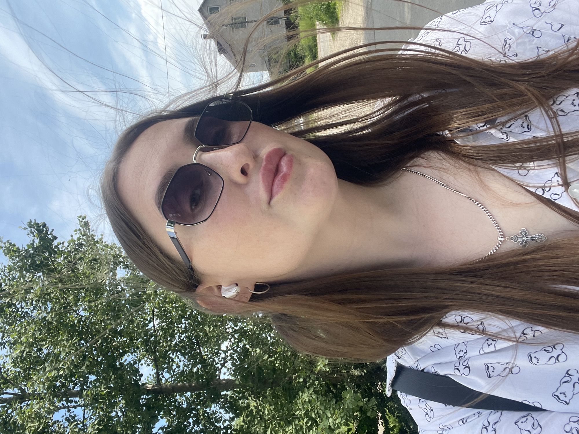 The image presents a head-and-shoulders view of a young woman in an outdoor environment, identified as Сырково, Russia. She is positioned centrally, gazing directly at the camera with a slight pout. Her long, straight brown hair is loose, with strands visibly blown by the wind, particularly to the right. She wears dark-lensed, octagonal sunglasses with silver-toned accents. A white earbud is inserted into her visible left ear, accompanied by a small silver hoop earring. She is dressed in a light-colored, collared shirt patterned with black outline figures resembling bears. A silver-toned curb chain necklace with a crucifix pendant hangs around her neck. A black strap, likely from a bag, is visible over her left shoulder. The background to the left features a large tree with dense green foliage against a bright blue sky. To the right, in the mid-distance, an aged, multi-story building with light-colored siding and a dark roof is visible, along with a dirt or unpaved path and sparse green vegetation. The scene is illuminated by bright daylight.