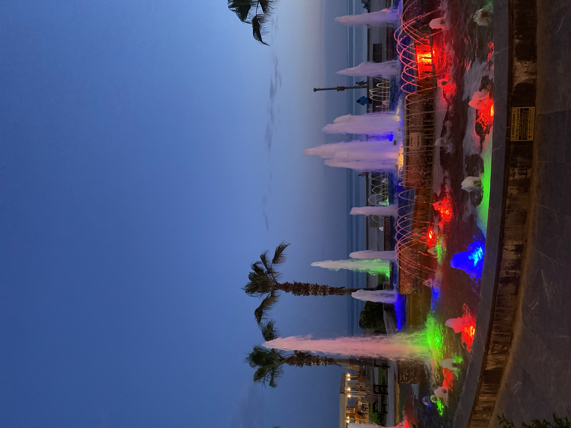This image captures an evening scene along a coastal promenade in Alanya, Turkey, featuring a lively, illuminated fountain display. The sky, a deep and rich blue, indicates dusk or nighttime, with faint, elongated cloud formations streaking across it.
In the midground, a long, rectangular water fountain is the focal point, with numerous jets of water shooting upwards. These water streams are vividly lit by an array of colorful lights in red, green, blue, and pink/purple hues, creating a dynamic and enchanting spectacle. Several tall palm trees are silhouetted against the dusky sky, primarily on the left side of the fountain, enhancing the tropical ambiance.
Beyond the fountain, a calm body of water, likely the sea, extends towards the horizon. In the distance, various indistinct structures and subtle artificial lights suggest a bustling coastline. A tall light pole stands behind the fountain, and a few blurred figures, possibly people strolling along the promenade, can be discerned near the water's edge.
On the right side of the fountain, a yellow warning sign is clearly visible. The text, in Turkish, reads "HAVUZA GİRMEK TEHLİKELİ VE YASAKTIR" (Entering the pool is dangerous and forbidden), followed by "ALANYA BELEDİYESİ" (Alanya Municipality), indicating public safety regulations. The scene conveys a peaceful and attractive atmosphere, with the colorful fountains serving as a beautiful evening attraction.