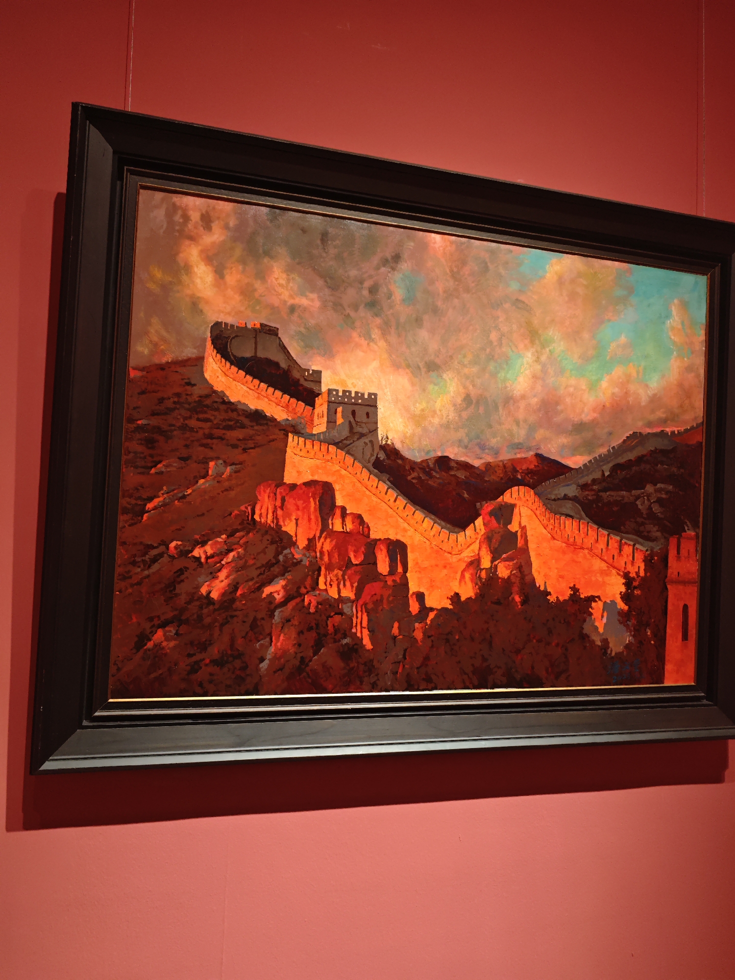 A framed painting of the Great Wall of China hangs on a red wall. The painting is primarily orange and red, with some blue and grey in the sky. The wall in the painting winds over a mountainous landscape. The location of the painting is unknown.