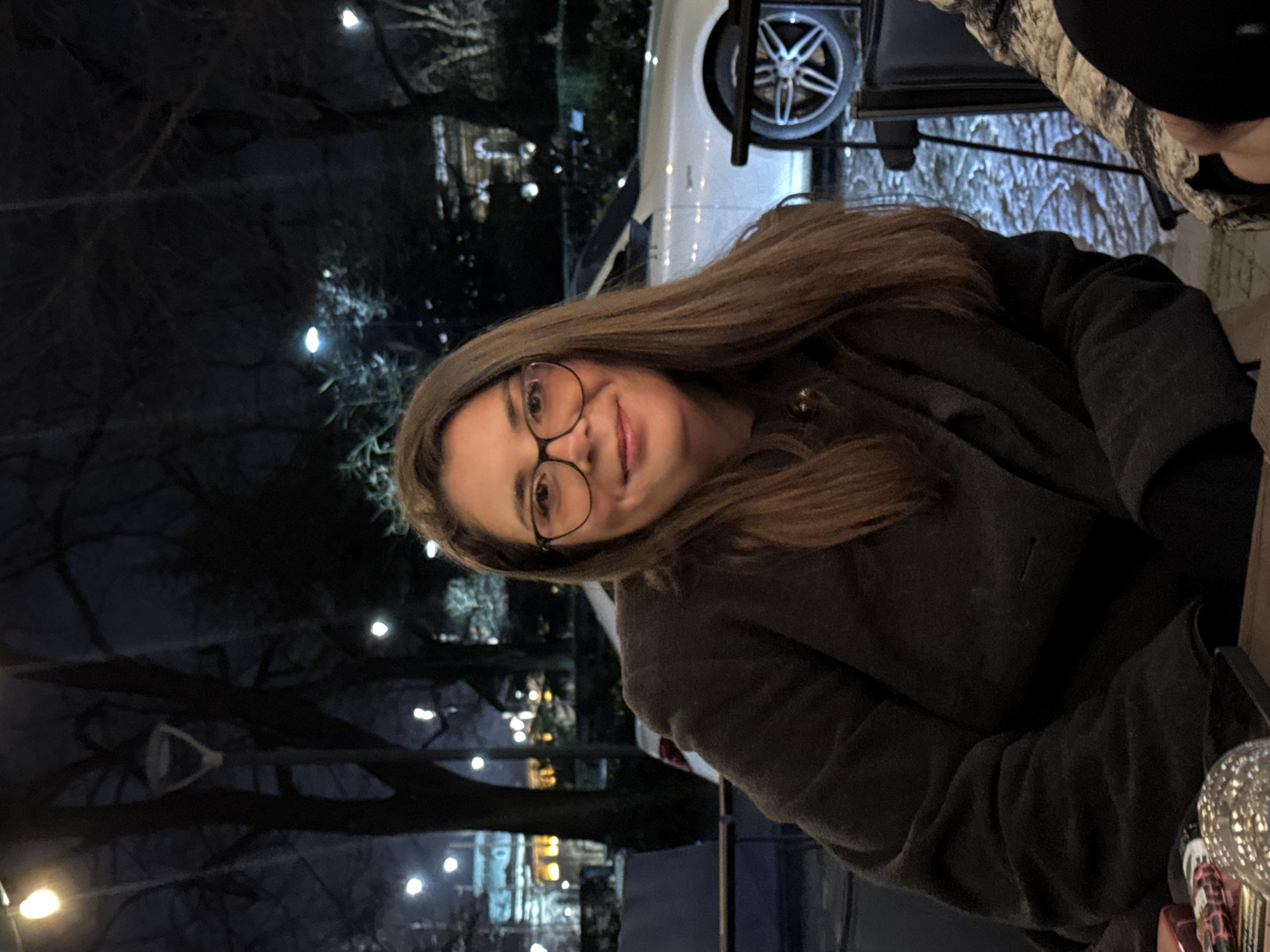 The image displays a female individual seated outdoors at night, looking directly at the camera with a slight smile. The subject is a Caucasian female with long, straight, brown hair, wearing black-rimmed glasses and a dark grey coat. She is positioned in front of a table, on which a red object and a clear, textured glass object are partially visible in the foreground left. A portion of a second individual's leg or arm, clad in camouflage-patterned fabric, is visible in the lower right corner. The background reveals an urban environment with cobblestone pavement. A white Mercedes-Benz vehicle, specifically the passenger side with a multi-spoke wheel, is partially visible behind the subject. Numerous spherical, warm-toned lights illuminate the scene, some appearing as strung lights, others as streetlamps, including a distinct bell-shaped streetlamp in the upper left. Bare tree branches are silhouetted against the dark sky. In the far left background, a large, ornate building with illuminated windows and classical architectural features is visible. The scene is consistent with an outdoor dining area or public space within a city at night.