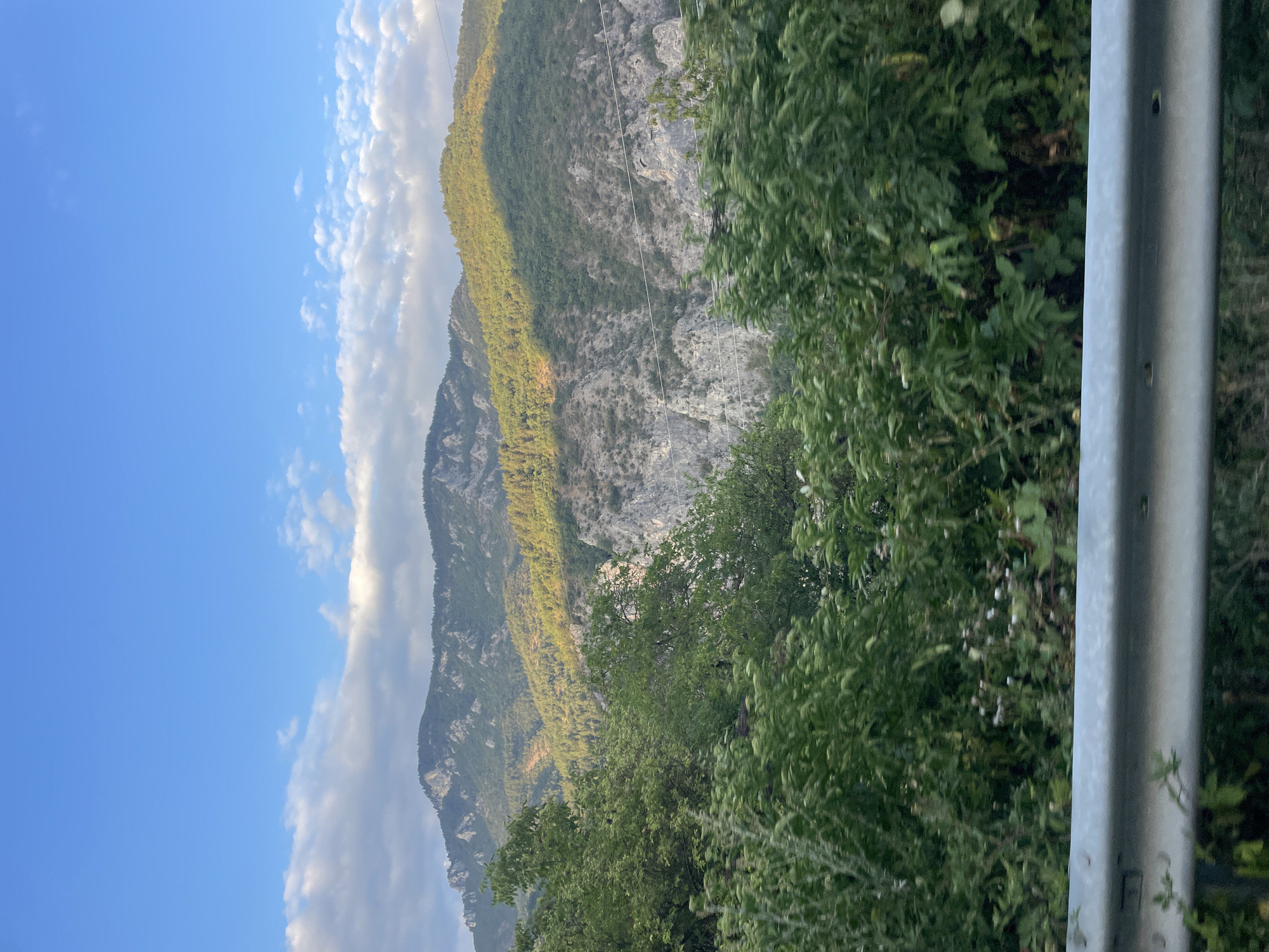 The image shows a mountainous landscape with a clear blue sky and scattered clouds. In the foreground, lush green foliage and trees are visible, with a silver guardrail running horizontally across the bottom of the frame. The mountains in the background are covered with dense forest, with some rocky outcrops and a deep gorge. Sunlight casts dramatic shadows across the mountainside, highlighting the contours of the terrain. The scene suggests a natural, undeveloped area, likely a natural park or a rural landscape. There are no people or any signs of urban activity. The scene is likely captured from a moving vehicle due to the slight blur in the foreground foliage. This view is consistent with the mountainous scenery found near Yugovo, Bulgaria, a village in the Rhodope Mountains known for its natural beauty and outdoor recreational opportunities.