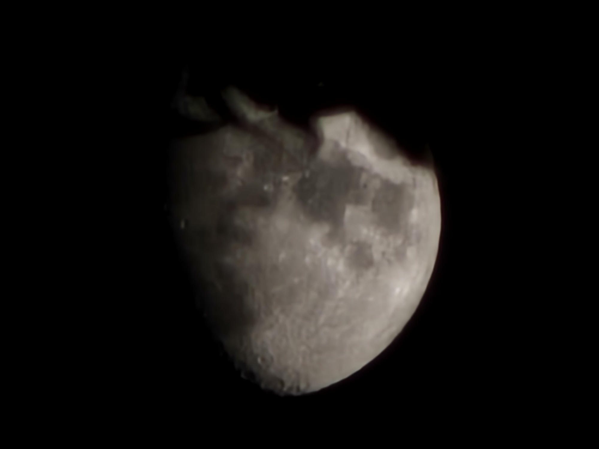 The image is a low-resolution, slightly blurry photograph of the moon against a black sky. The moon is partially illuminated, showing craters and surface features. A dark, irregularly shaped shadow or object obscures a portion of the moon's right side. The image does not contain any elements that would allow for identification of a specific location, such as landmarks, buildings, or cityscapes. Therefore, it is impossible to determine if this image was taken in Denpasar, Indonesia, or any other specific city.