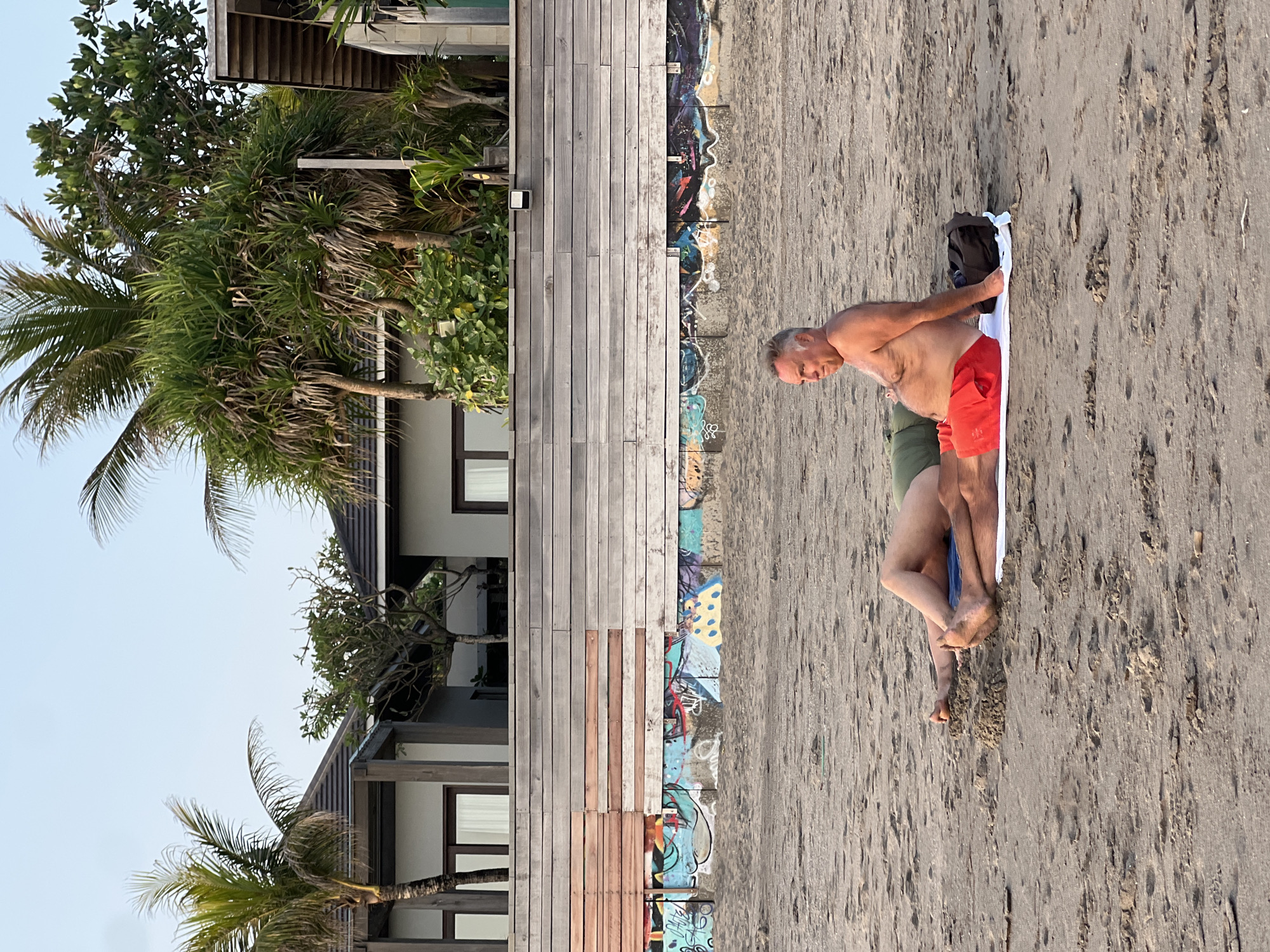 Two men are lying on a sandy beach in North Kuta, Indonesia. The man in the foreground, wearing red swim trunks, is sitting up and looking to the left. The man behind him, wearing green shorts, is lying down. They are both on a white towel. In the background, there is a wooden wall with graffiti and several palm trees. Beyond that, there are modern buildings with flat roofs. The sky is clear and light blue.