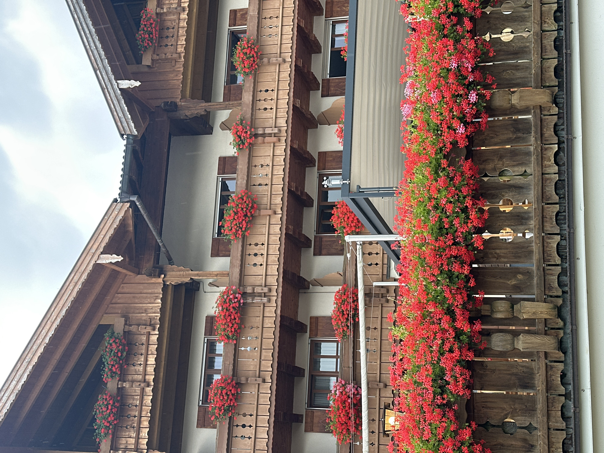The image shows a multi-story building in Seeon-Seebruck, Germany, decorated with numerous planters filled with vibrant red flowers. The building features traditional alpine architecture with extensive use of wood for balconies, eaves, and decorative elements. The balconies are adorned with railings and filled with overflowing planters. Windows with wooden shutters are visible on each level. A wooden fence with carved details runs along the bottom of the image, also lined with red flowers. The sky is overcast, suggesting cloudy or rainy weather. The scene depicts a residential or hotel building, possibly in a tourist area, emphasizing a charming and picturesque aesthetic with abundant floral displays.