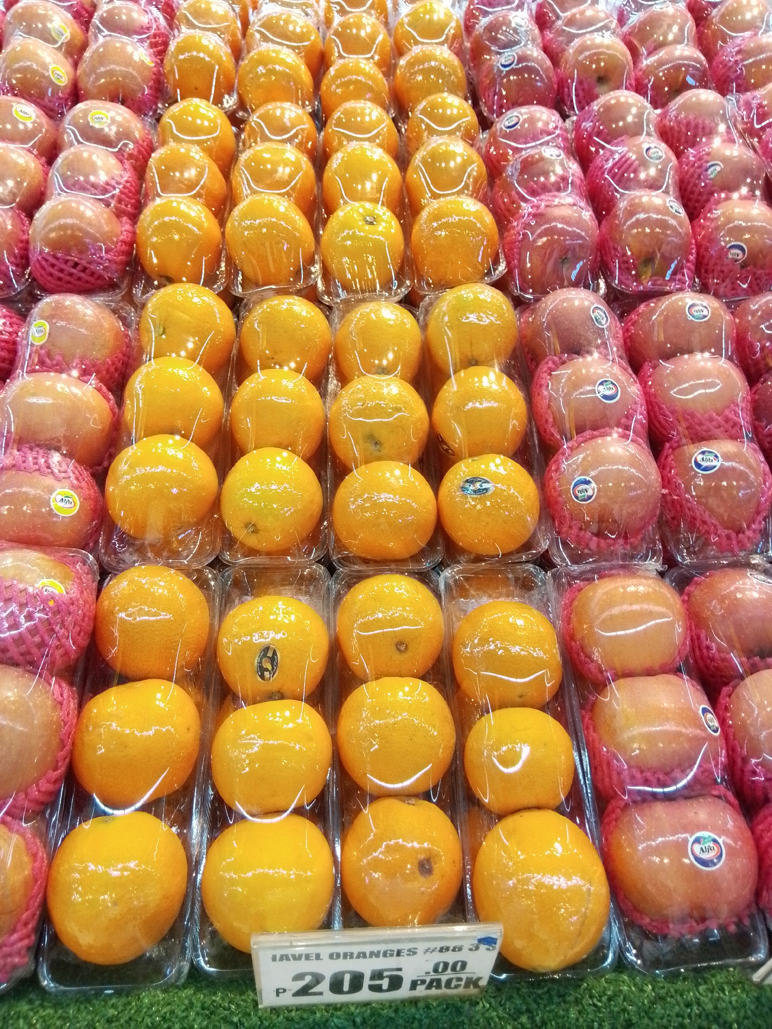 This is a close-up, eye-level, medium shot of a display of packaged fruits, likely in a market or grocery store. The fruits are primarily oranges and reddish-pink fruits, possibly apples or pears, presented in clear plastic packaging with some of the reddish-pink fruits also wrapped in pink foam netting. The display is arranged in rows and columns, with each fruit individually contained within a clear plastic tray. Some of the oranges have small circular stickers with blue and white designs, and some of the reddish-pink fruits have stickers with the brand name "Alfa" and a blue and white logo. A price tag is visible in the foreground, indicating "JAVEL ORANGES #B8 3'S P-205 .00 PACK". The background is blurred but appears to be a continuation of the fruit display. The lighting is bright and even, suggesting an indoor setting. The overall impression is one of fresh produce neatly presented for sale. The presence of the "JAVEL ORANGES" price tag suggests this scene could be in the city of Davao City, Philippines, known for its fruit markets.