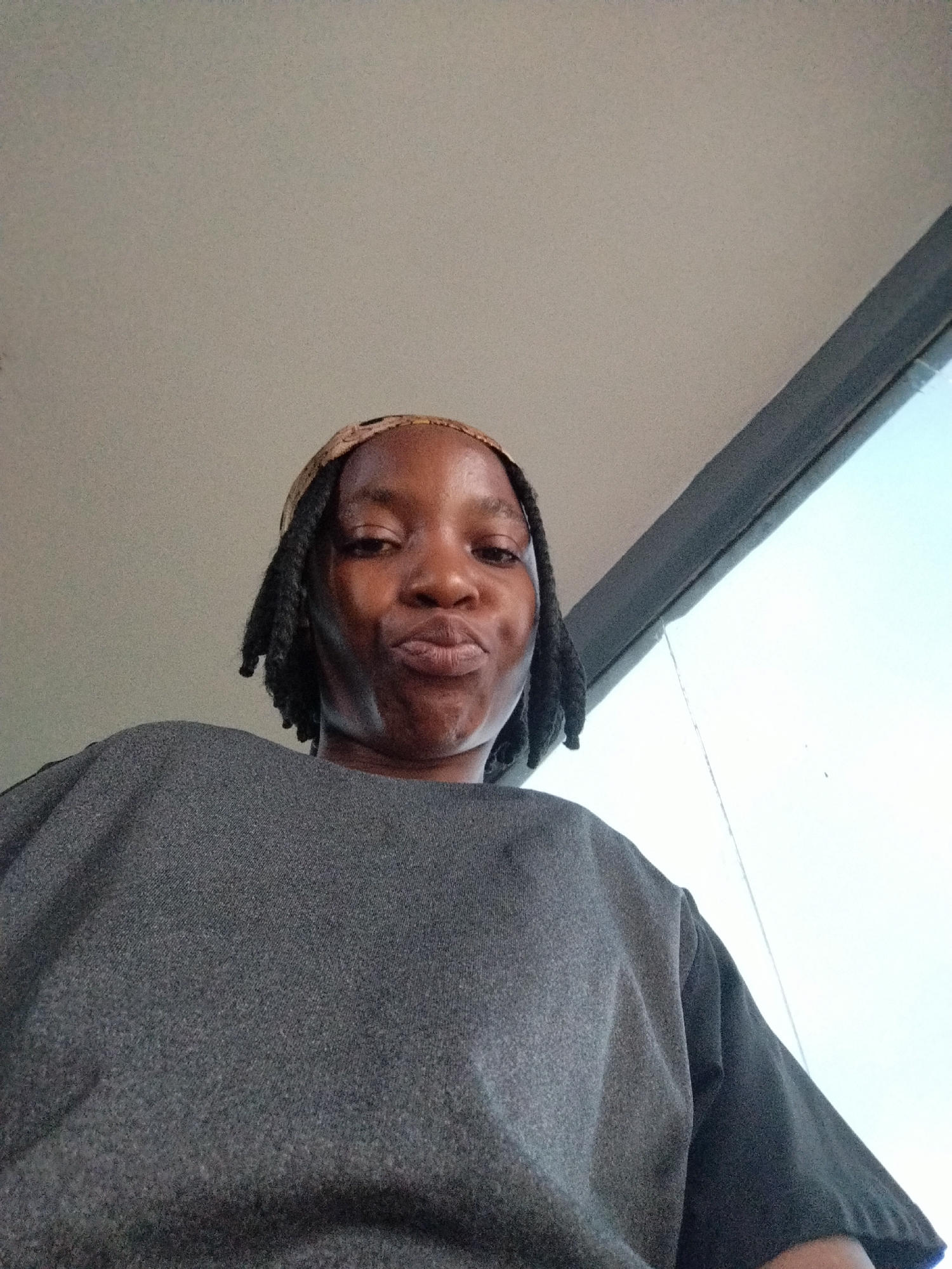 In Uselu, Nigeria, a medium shot captures a woman with dark skin and braided hair, wearing a dark grey shirt and a patterned headband. She is positioned in the foreground, looking directly at the camera with her lips pursed. The background consists of a light-colored ceiling or wall and a large window with a bright sky visible through it, suggesting an indoor location with natural light. The perspective is from a low angle, looking up at the woman.