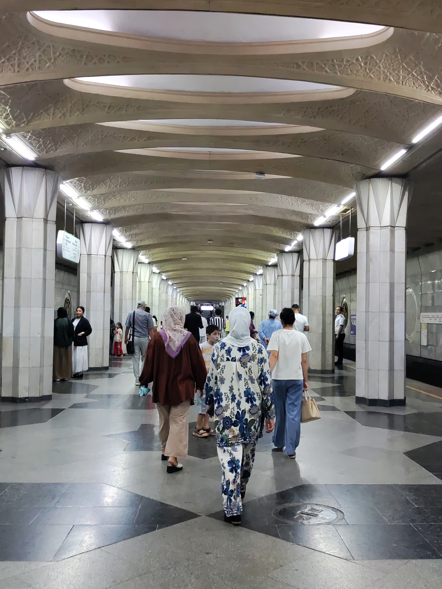 The image depicts the interior of the Tashkent Metro, specifically the Kosmonavtlar station. The station features a distinctive architectural design with a vaulted ceiling adorned with geometric patterns and recessed lighting. Rows of fluted columns, painted white and grey, support the ceiling, creating a sense of grandeur and depth. The floor is a patterned mosaic of dark and light grey tiles.
Several individuals are visible walking through the station. In the foreground, a woman wearing a white outfit with a blue floral print is walking away from the camera. To her left, a woman in a dark brown jacket and light-colored pants, also wearing a headscarf, is walking in the same direction. Further into the station, other people are seen walking, some in groups and others individually. Two women in traditional attire, one in a dark green abaya and the other in a white dress with a black cardigan, stand near a column on the left. A young girl in a red dress is visible in the middle ground. A man in a white t-shirt and blue pants is walking towards the foreground, carrying a beige bag. On the far right, a man in a white shirt and black pants stands facing the camera. Signage is visible on the walls and hanging from the ceiling, indicating the station's identity. The overall atmosphere is that of a busy public transit space with a unique, ornate aesthetic.
