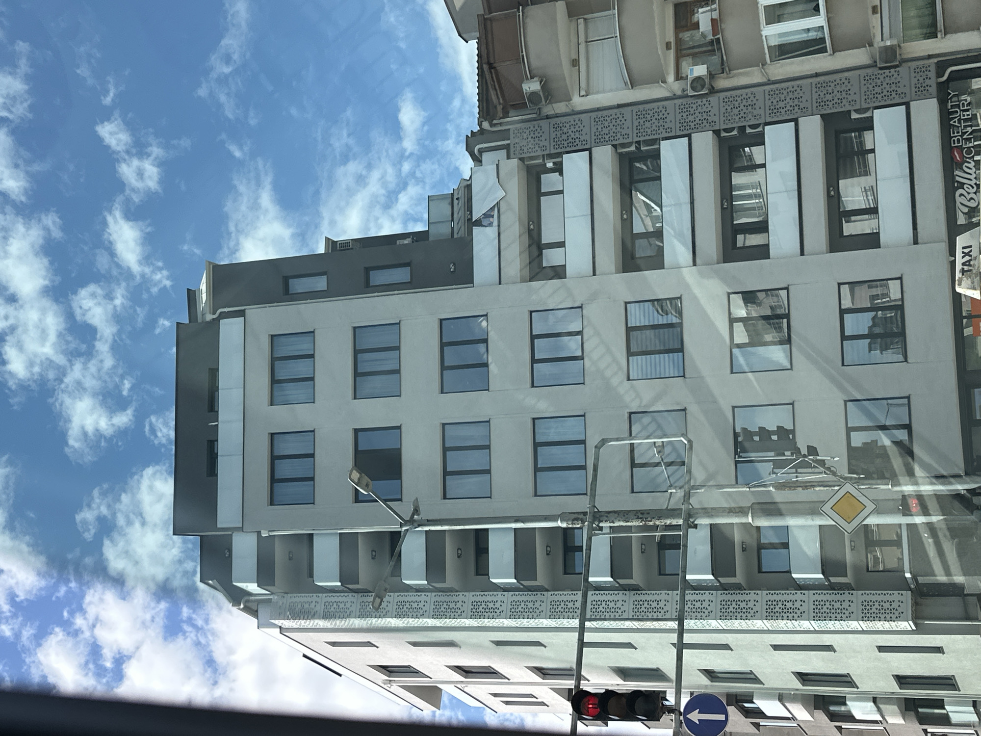 The image depicts a view of a modern building in Plovdiv, Bulgaria, from a low angle, possibly from inside a vehicle. The building has a light-colored facade with multiple windows and a geometric design on some floors. A sign "Bella Beauty Center" is visible on the side of the building. In the foreground, a street light with a diamond-shaped yellow traffic sign and traffic lights is visible. The sky is blue with scattered clouds.