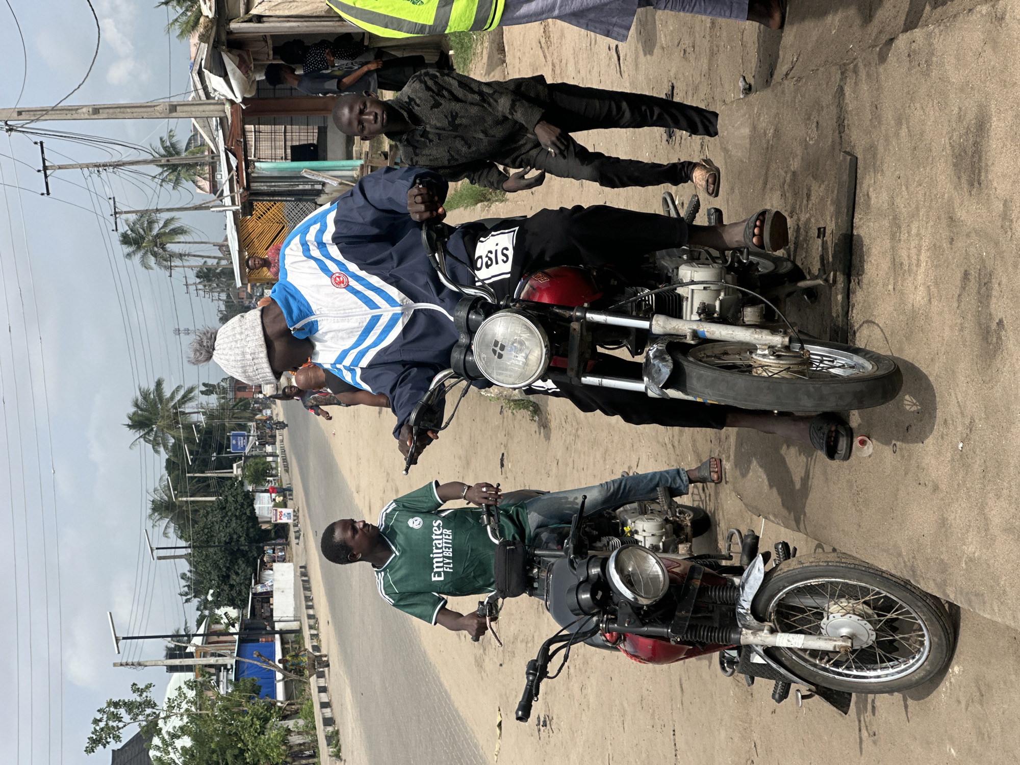 In Badagry, Nigeria, a street scene unfolds with several people and motorcycles. In the foreground, a young man wearing a green jersey with "Emirates Fly Better" visible is seated on a red motorcycle. To his right, another young man in a blue and white jacket with a gray beanie is also on a motorcycle, looking to his left. Between the two motorcycles, a younger boy in a camouflage jacket stands and looks towards the viewer. The background reveals a dusty road with more people, structures that appear to be small shops or homes, palm trees, and utility poles with numerous wires. The environment suggests a typical day in a bustling Nigerian town.