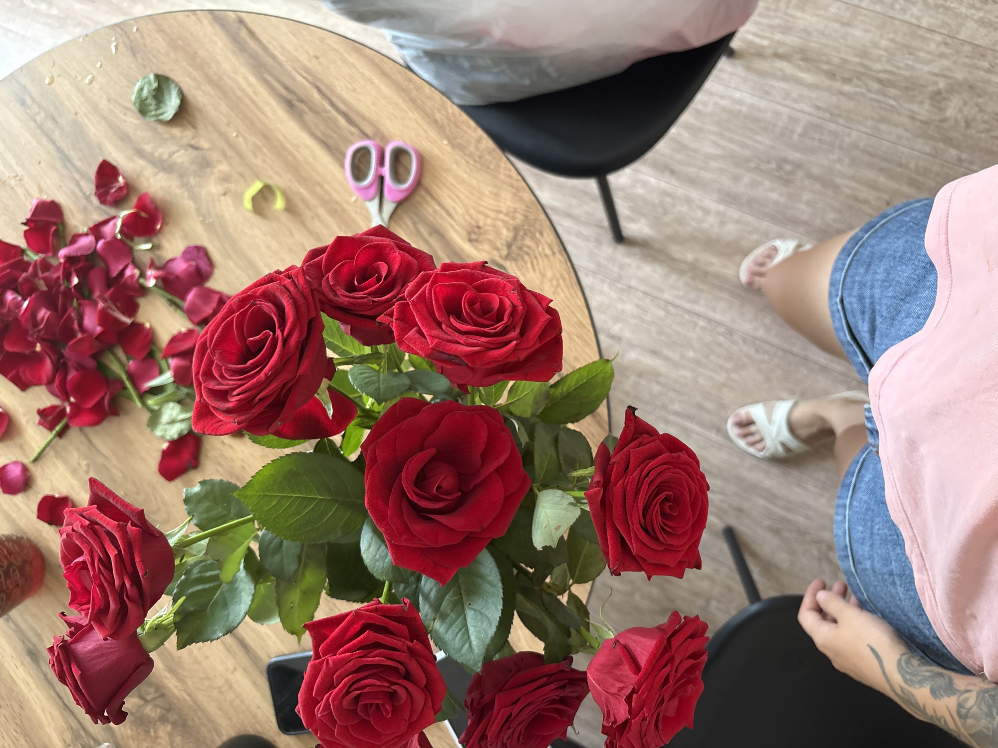 An overhead shot shows a bouquet of red roses arranged on a round wooden table in Rostov-on-Don, Russia. Scattered rose petals and a pair of pink-handled scissors lie on the table's surface. In the lower portion of the image, a person's legs and torso are visible, wearing denim shorts and a pink t-shirt, with white sandals on their feet. The person's left arm, adorned with a tattoo, is also visible. The setting appears to be indoors, with a wooden floor beneath the table and chair.